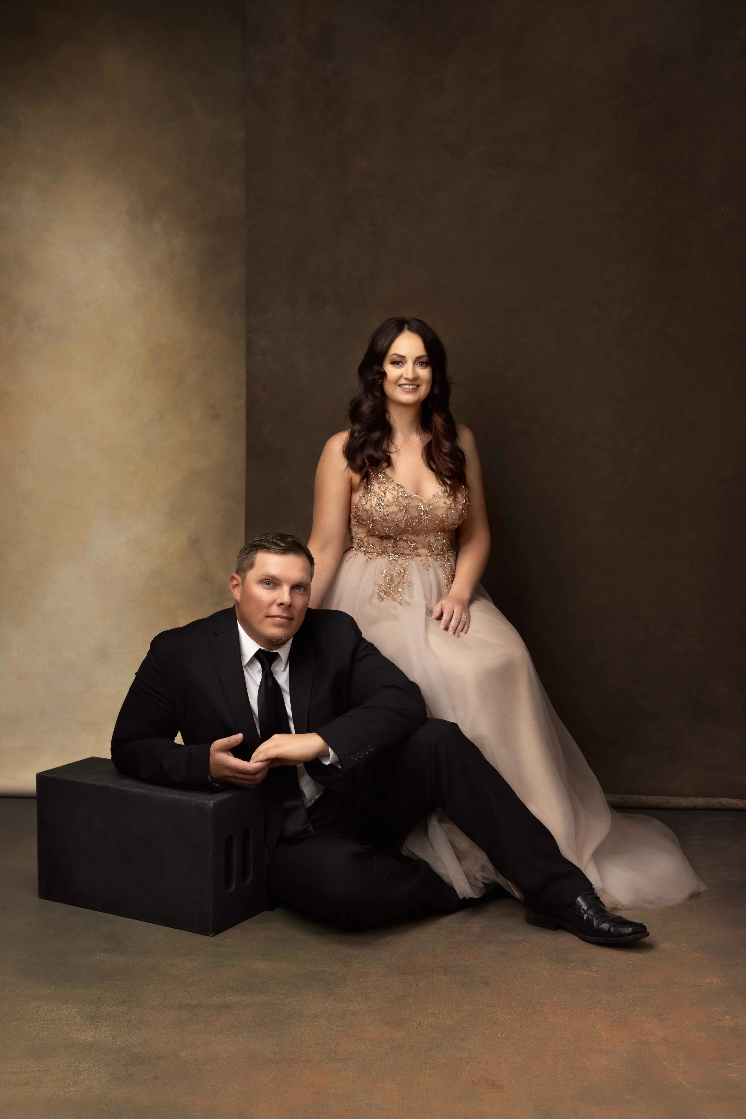 A man in a black tuxedo sitting with his hands clasped on his knee, and a woman in a beige formal gown with intricate embroidery, sitting behind him against a textured brown and yellow wall.