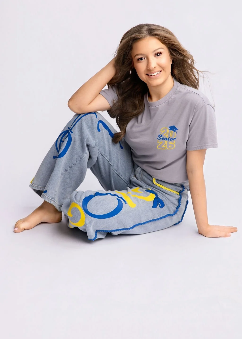 Young woman sitting barefoot on the floor, smiling, wearing a gray '2026 Senior' t-shirt with blue and yellow design, and jeans with blue and yellow accents.