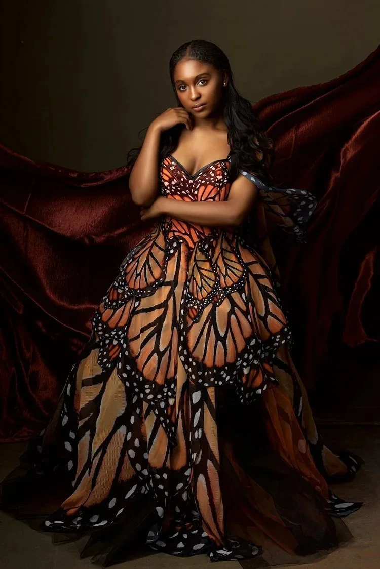 A woman in a butterfly-themed dress with butterfly wing patterns and colors standing against a dark background, posed with her hand near her chin and looking at the camera.