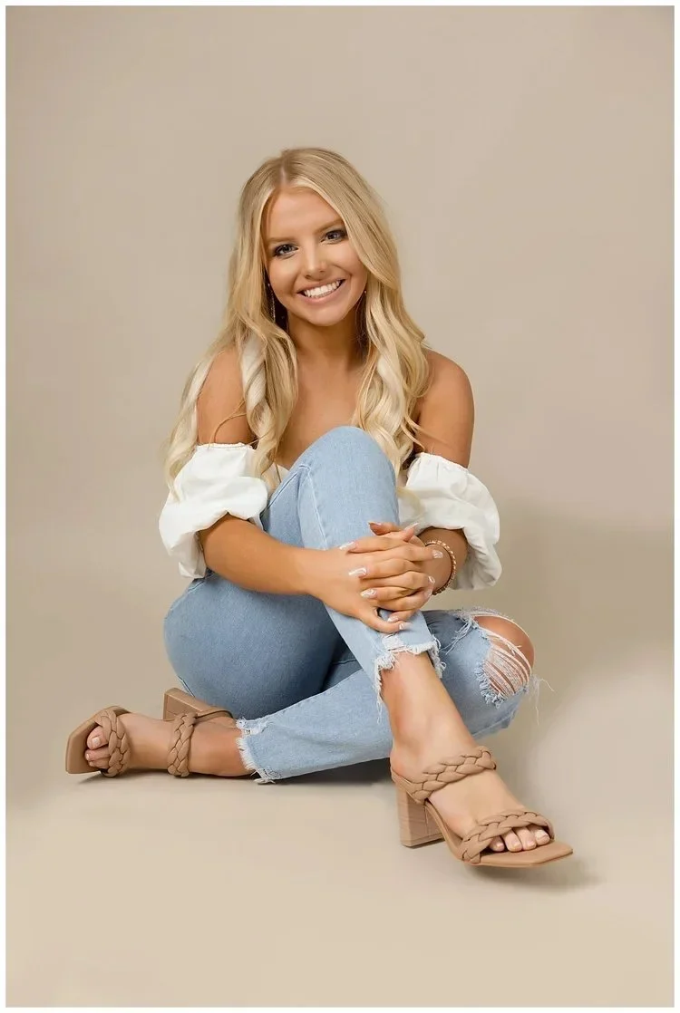 A young woman with long blonde hair, smiling, sitting on the floor against a plain beige background. She is wearing a white off-the-shoulder top, ripped light blue jeans, and beige braided heeled sandals.