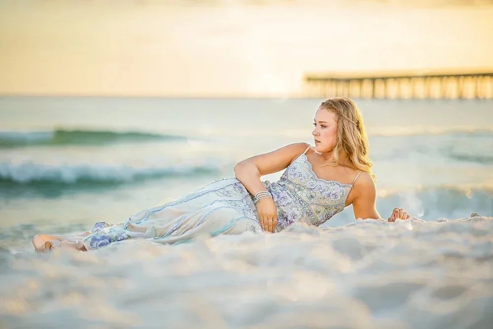 A woman in a detailed white dress lying on the sand at the beach with ocean waves and a pier in the background during sunset.