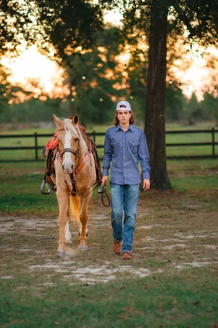 A young person with shoulder-length hair, wearing a white cap, blue polka dot shirt, and jeans, is walking a light-colored horse on a dirt path in a park-like setting during sunset.