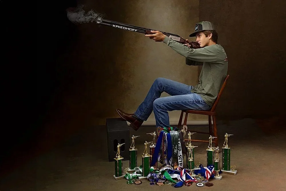 A young man sitting on a wooden chair aiming a rifle, with smoke coming from the barrel, surrounded by trophies and medals on the floor.