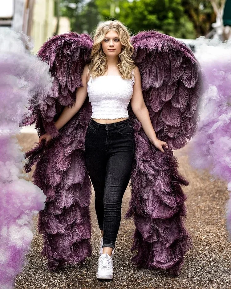 Young woman walking outdoors wearing large purple feathered wings costume with purple smoke around her.