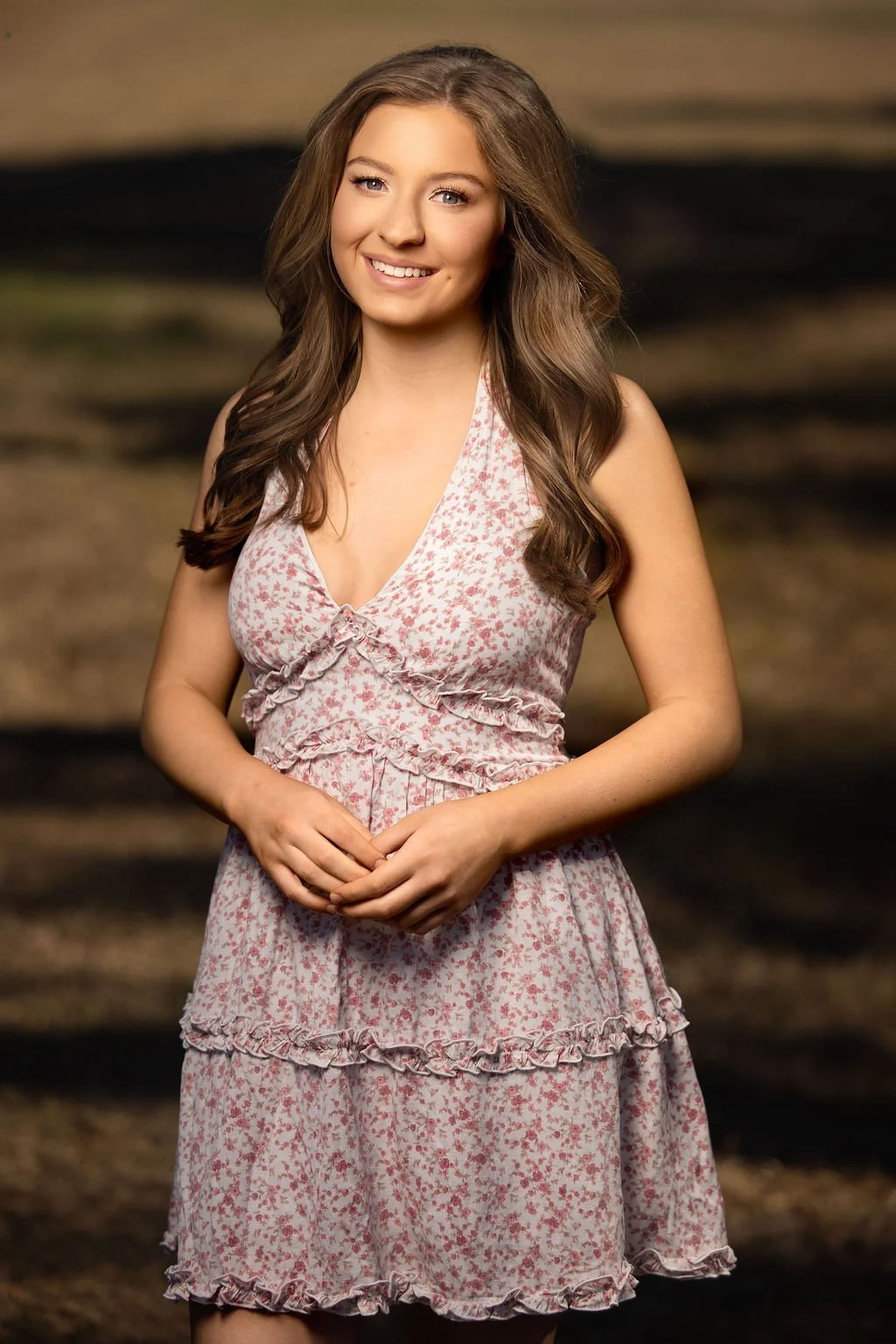 Young woman with long brown hair smiling at camera, wearing a pink and white floral dress, standing outdoors with blurred background.