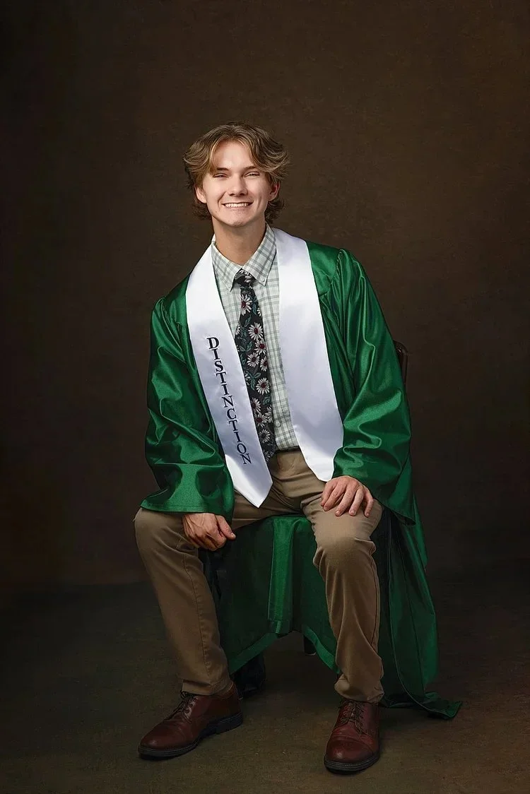 Young man in green graduation gown and white sash with 'DISTINCTION' written on it, sitting on a chair against a dark background, smiling.