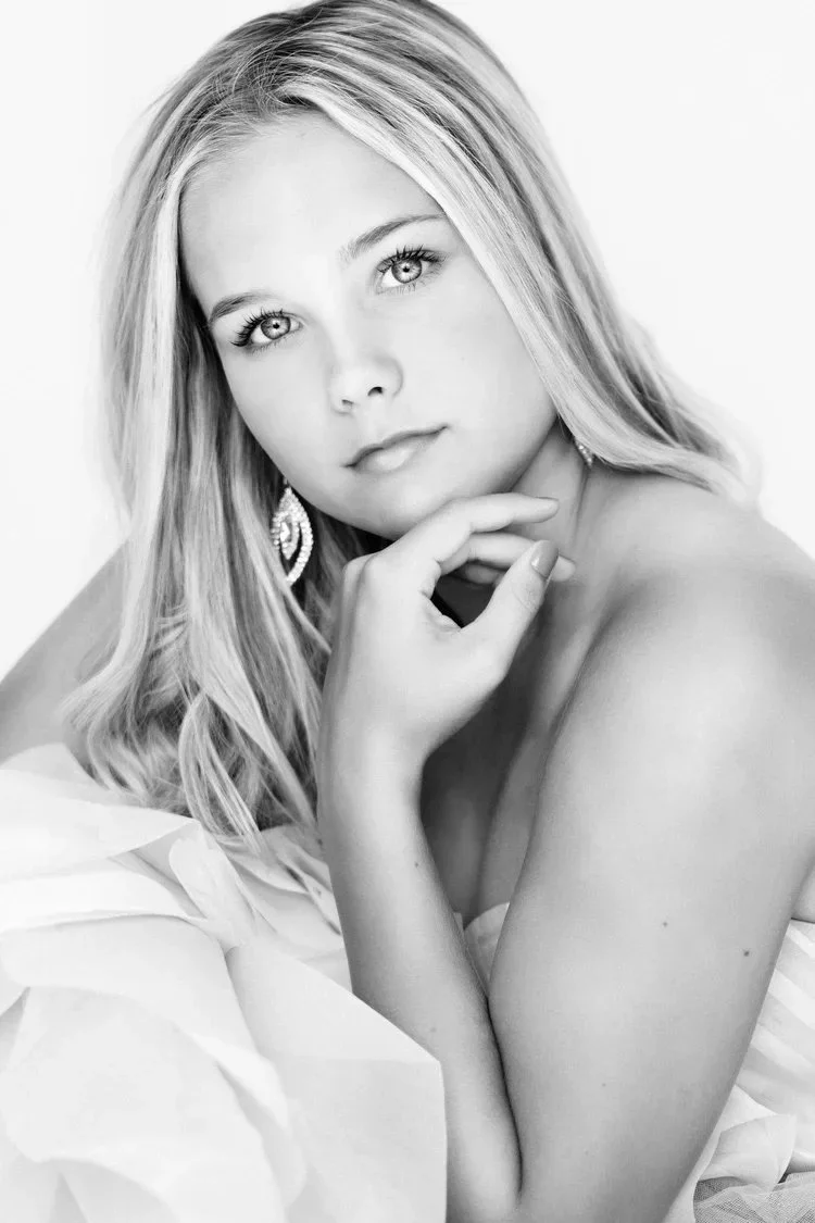 Black and white portrait of a young woman with long, wavy hair, looking into the camera with her chin resting on her hand, wearing elegant earrings, and draped in a soft fabric.