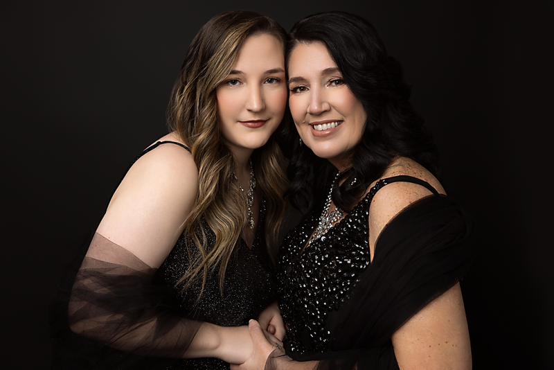 Mother and daughter dressed in black, smiling and holding hands against a dark background.