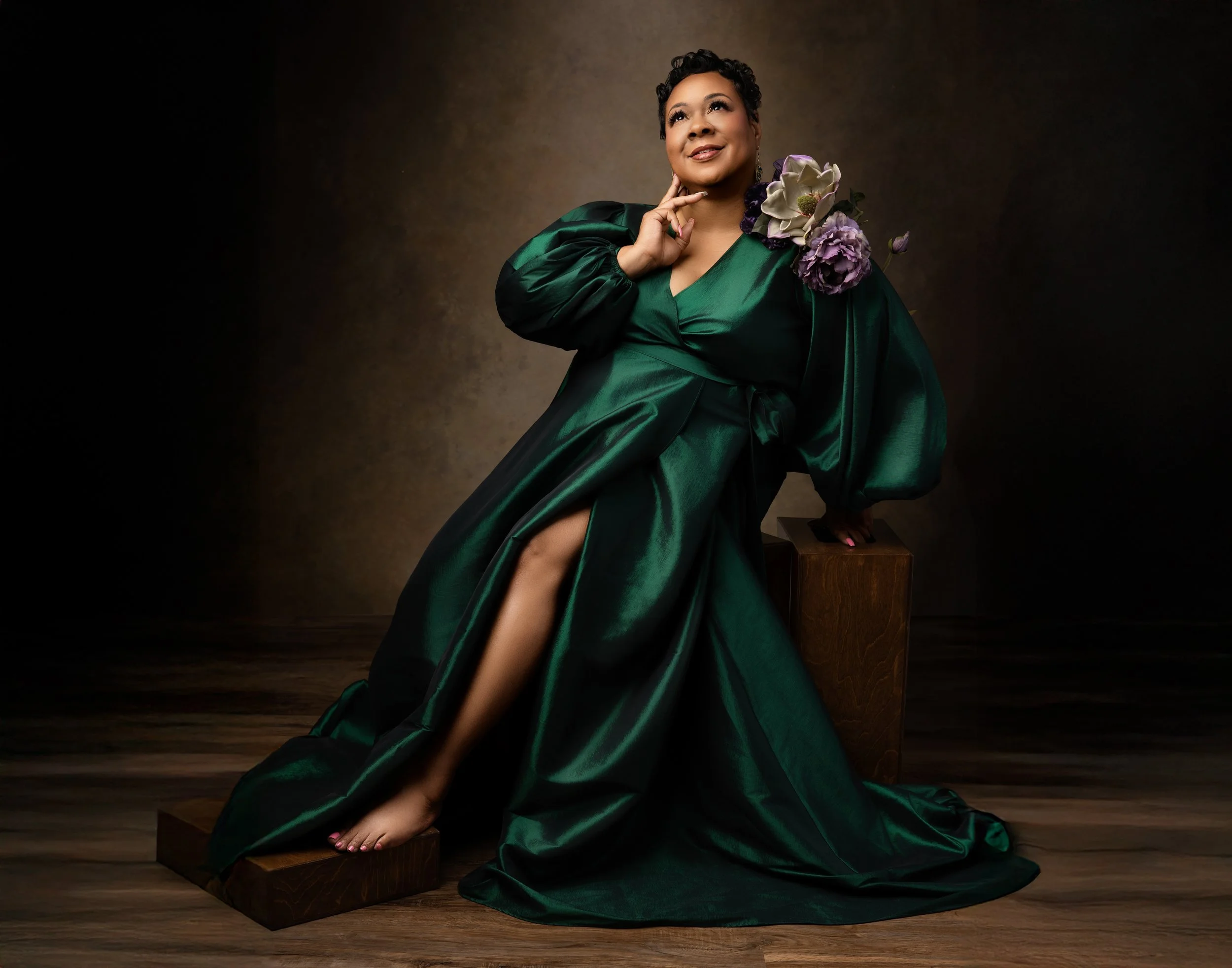 A woman wearing a dark green satin gown with a thigh-high slit, sitting on a wooden block, with a brown background. The gown has floral accents with large white and purple flowers on her shoulder, and she is touching her chin with her right hand, loo