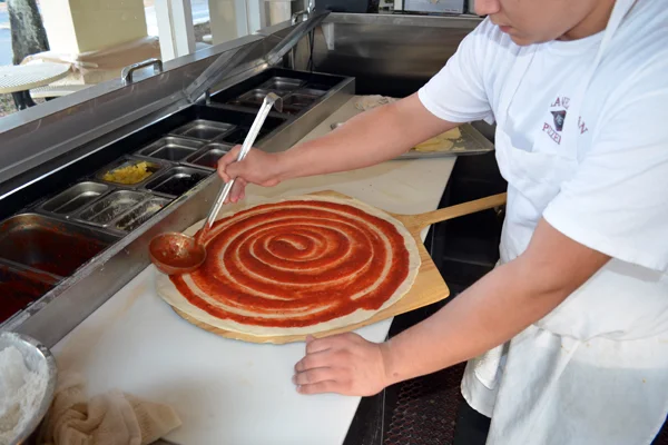 Pizza dough sitting on top of a large wood scapula being topped with spaghetti sauce in a circular motion, by a gentleman with a larger silver  spoon in a kitchen setting