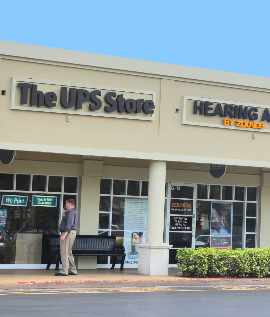 Exterior image on the UPS store,  with a man in black shirt and khaki pants standing in front for the store and under the store sign in black letter " The UPS Store"