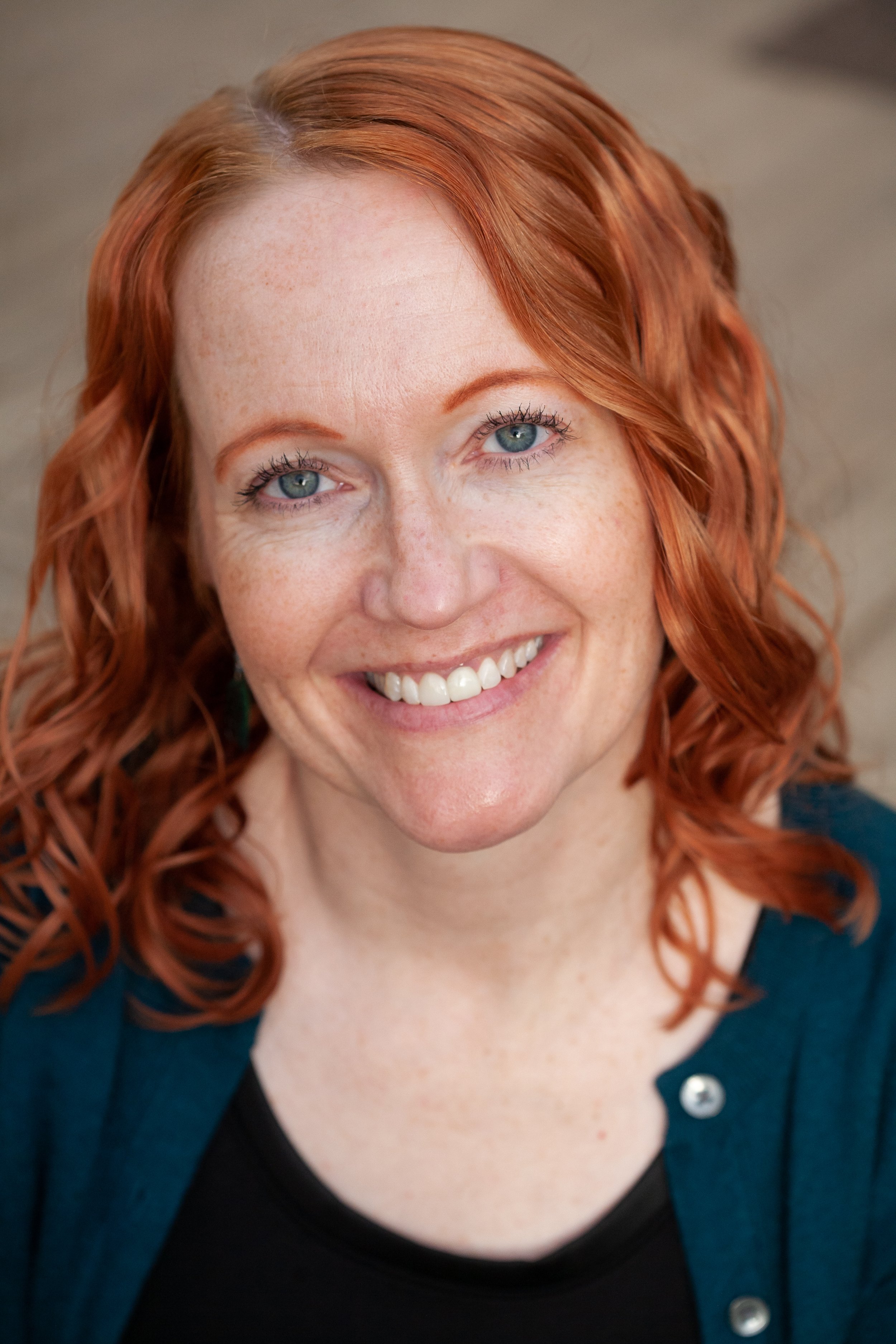 Photo of SLC Therapist Christy Kobe, LCSW, CCTP, EMDR Therapist Utah who has wavy red hair and blue eyes and is smiling, wearing a black blouse and a teal cardigan sweater