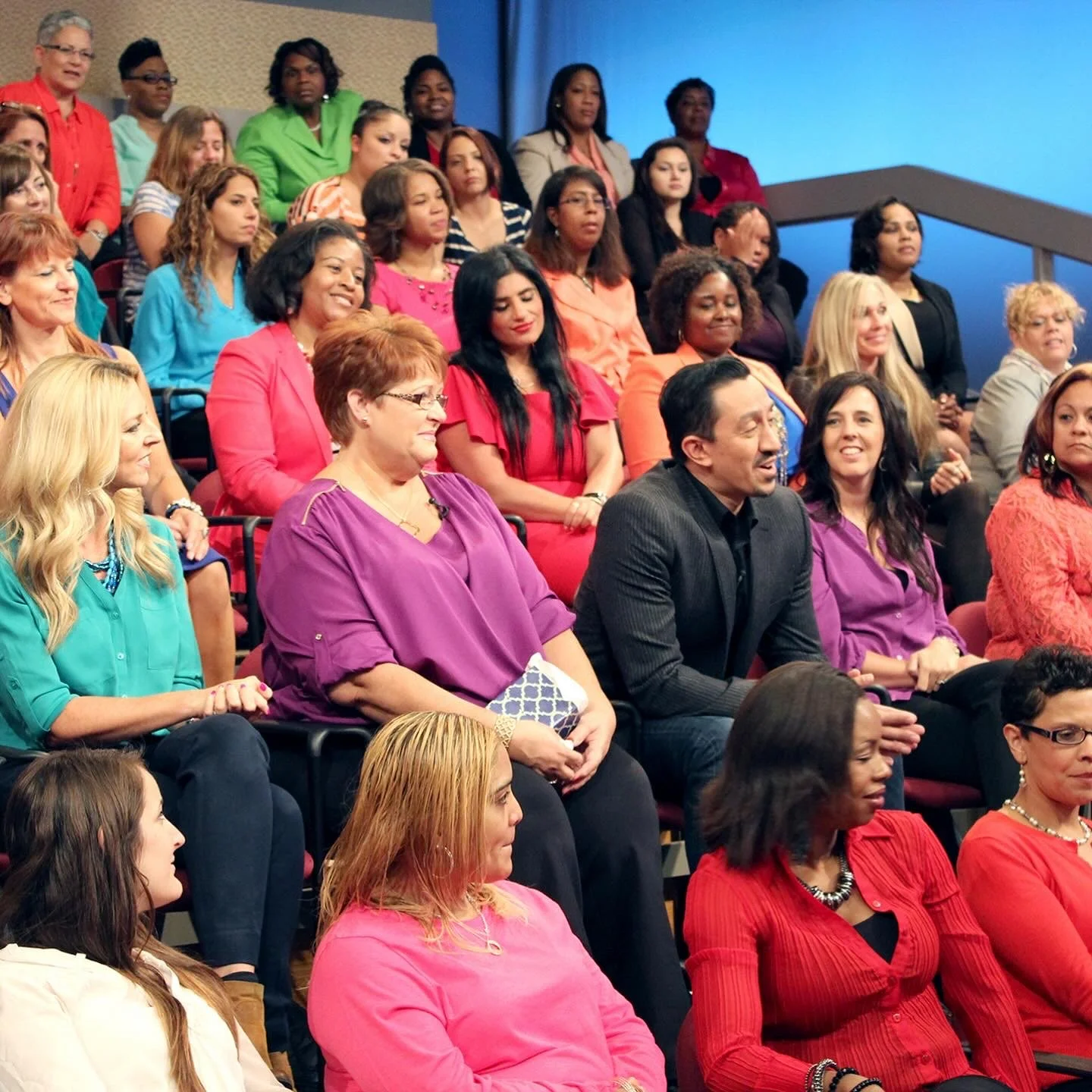 Several women are seated in attendance at The Steve Harvey Show. The audience is diverse, dressed in colorful clothing, and smiling during the event. Adam Mock sits in the middle of the audience speaking toward the main stage.