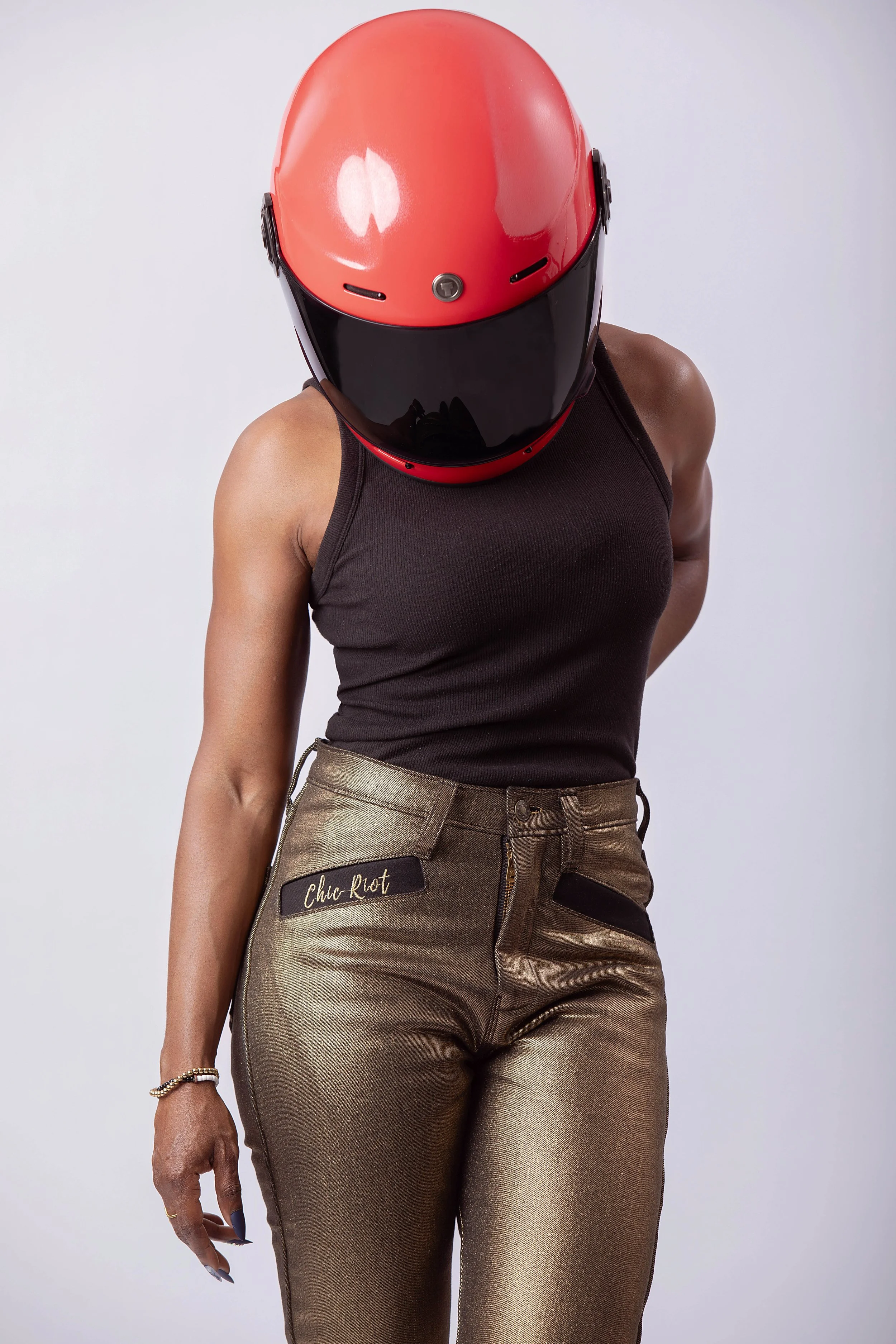 Model wearing metallic gold moto pants and black tank with red helmet, front view showing fit and styling
