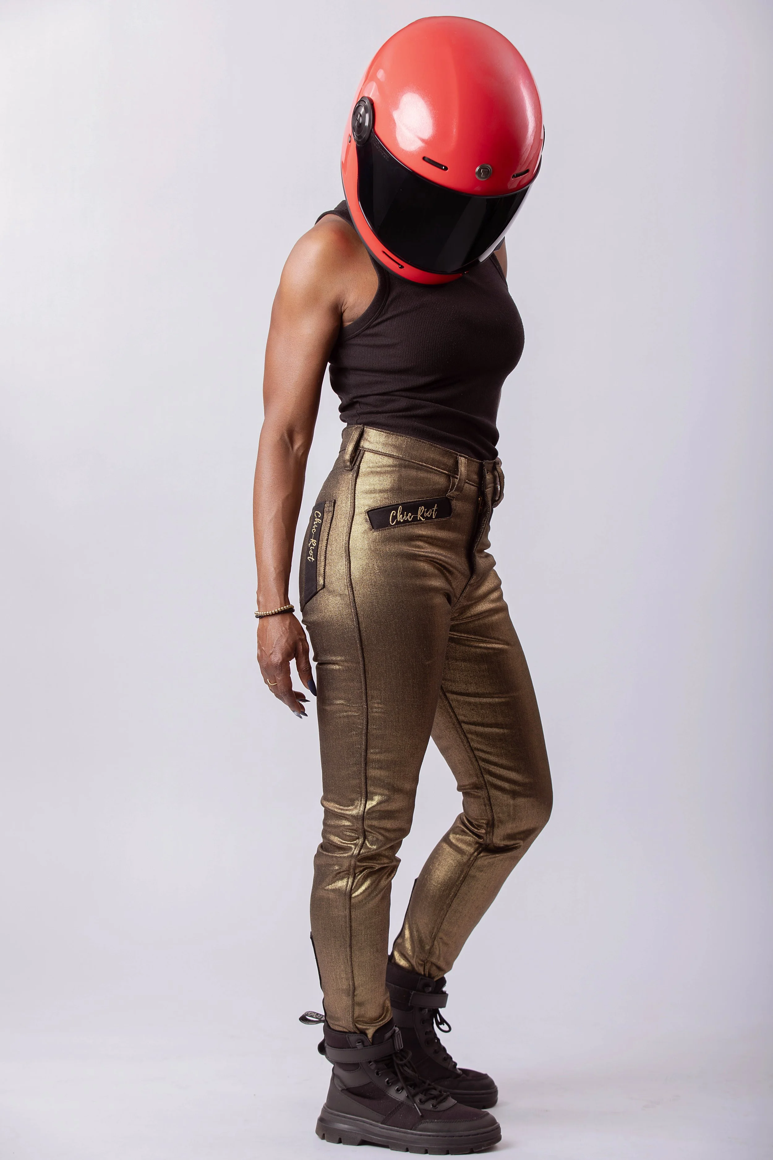 Model wearing metallic gold moto pants and black tank with red helmet, side view showing fit and styling