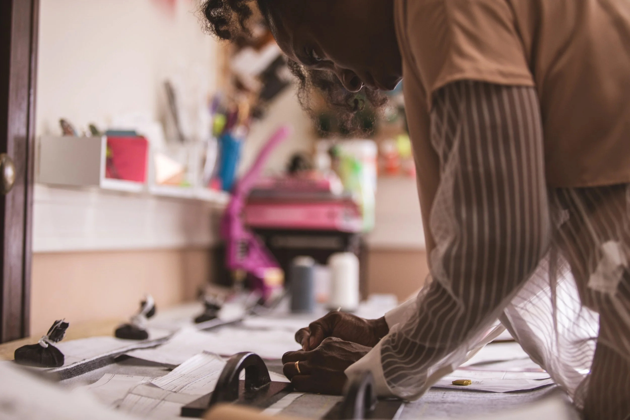 Woman wearing a brown shirt working on a sewing pattern at a table with black and white fabric and sewing materials. Shelves and various craft supplies are visible in the background.