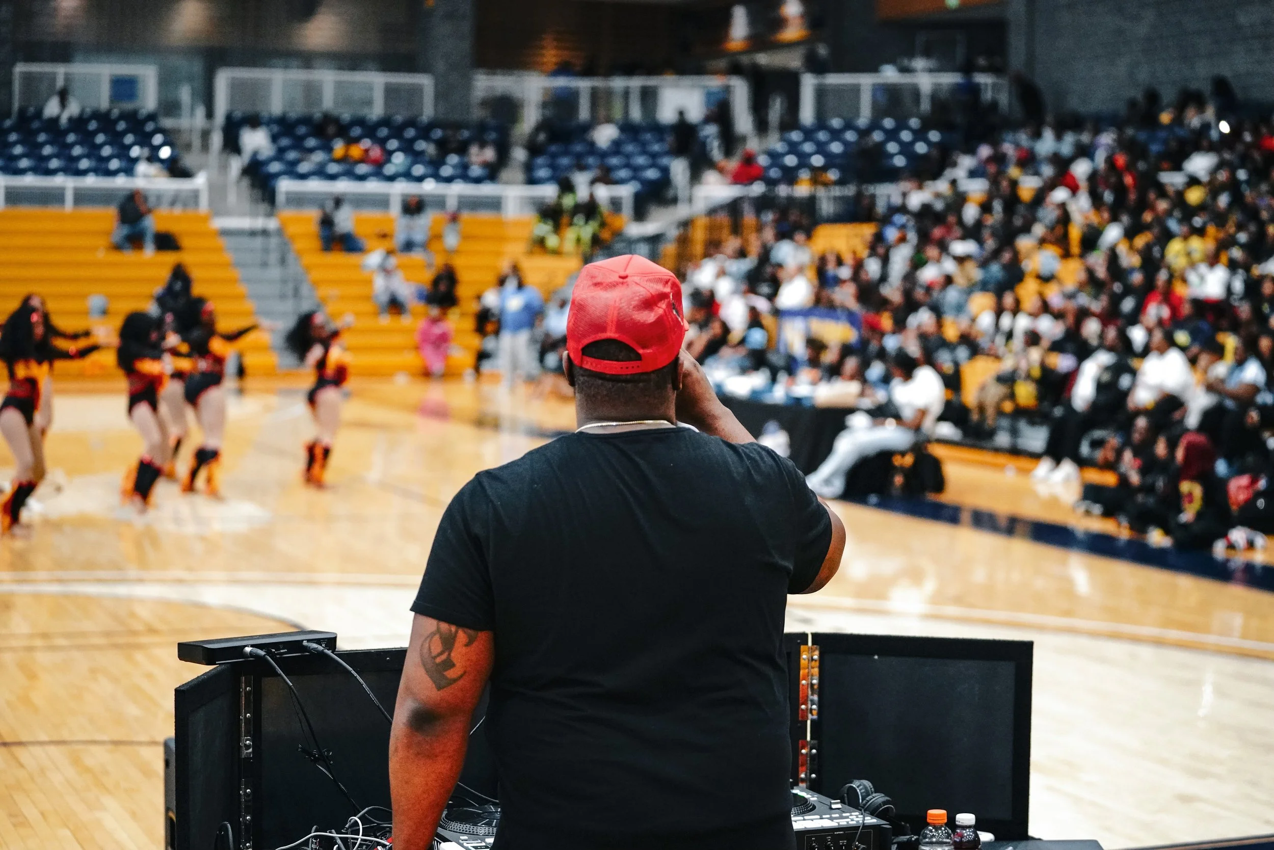 DJ Foqus wearing a red cap and black t-shirt stands behind DJ equipment in a gymnasium with a basketball court. In the background, a dance team in black and orange uniforms performs, and an audience is seated in the bleachers, watching the event.