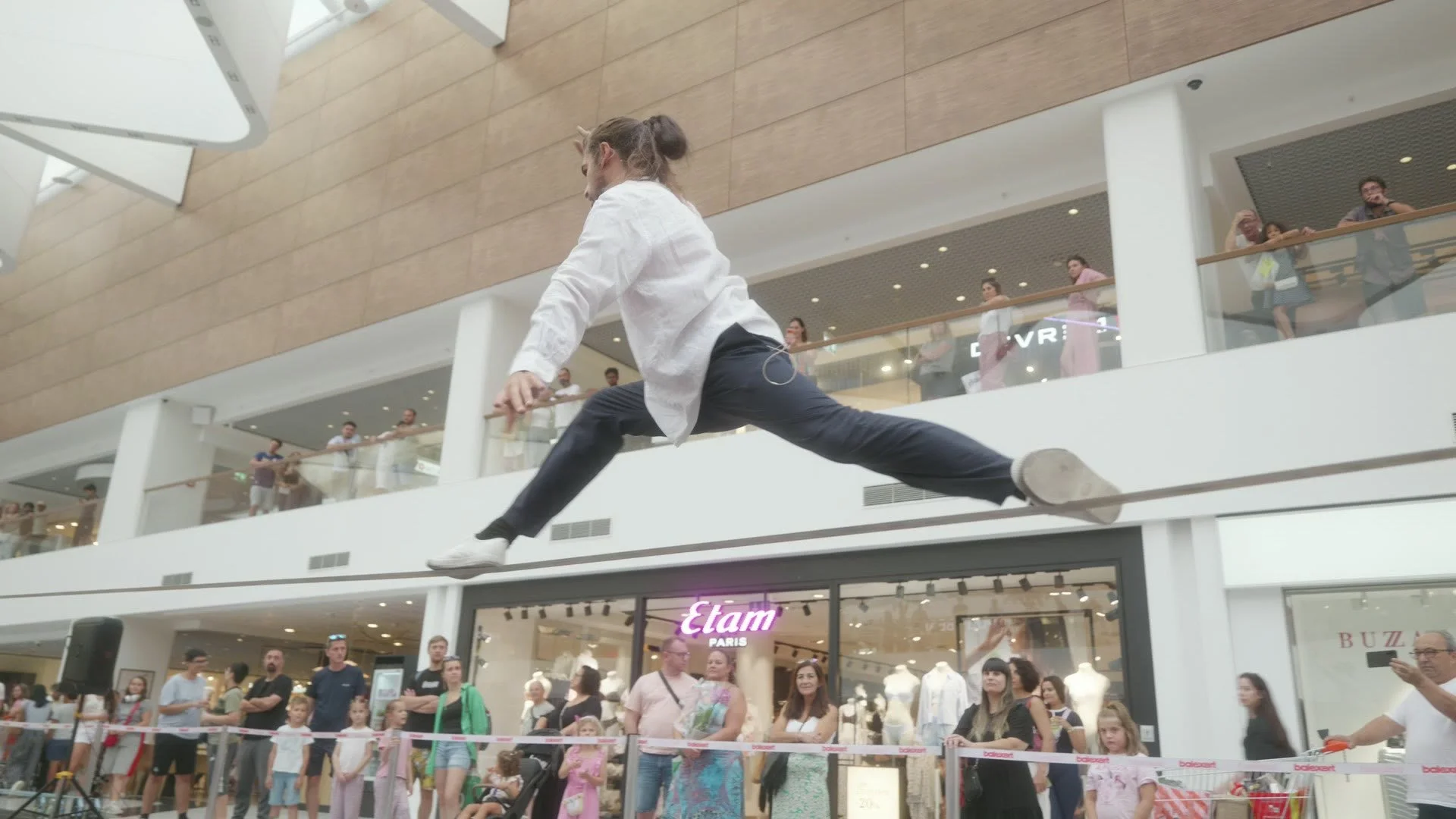 Artiste de slackline acrobatique en grand écart au-dessus de la ligne dans l'atrium du centre commercial Balexert à Genève, public au sol et aux étages