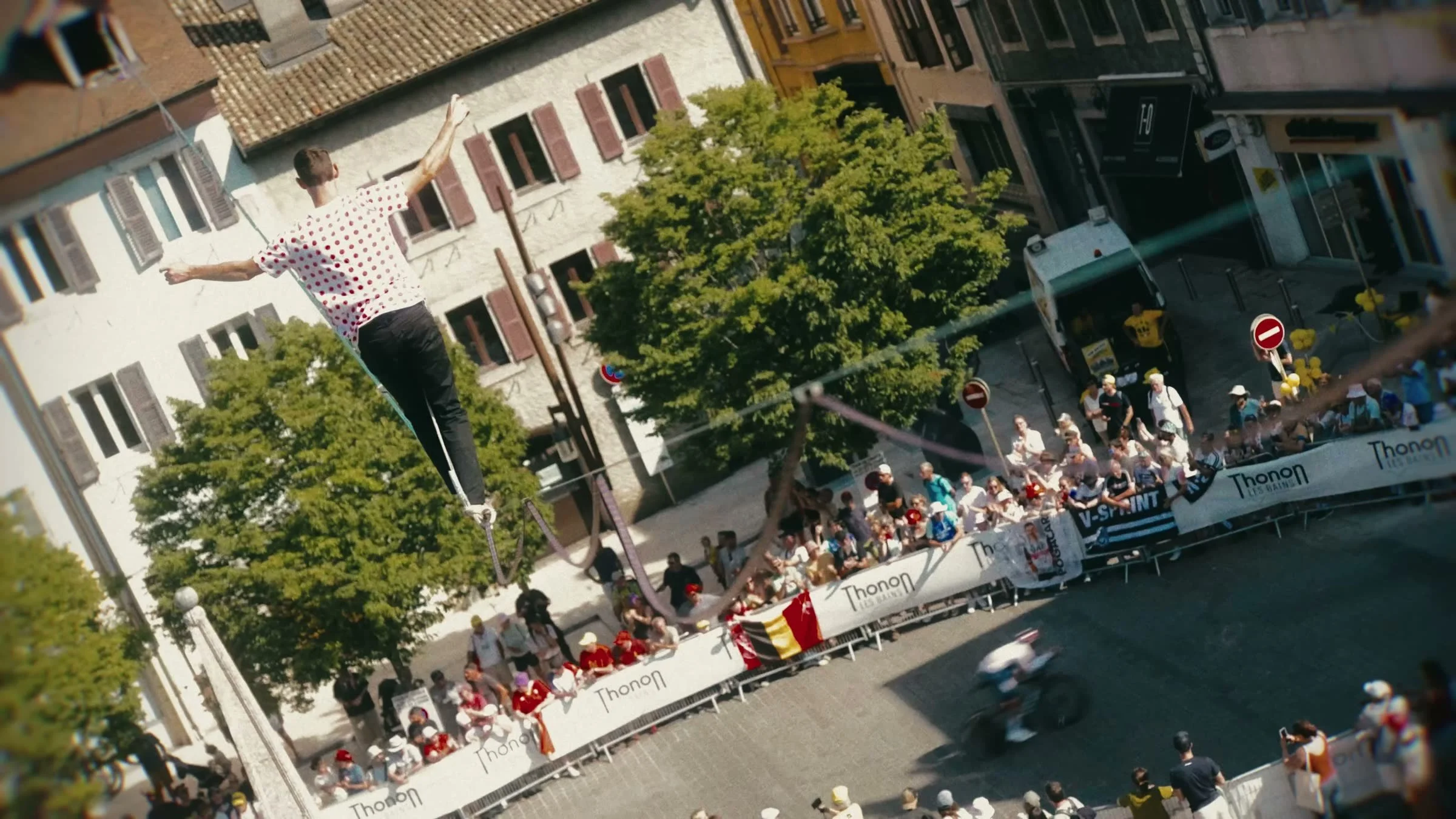 Artiste de highline en maillot à pois au-dessus du parcours du contre-la-montre du Tour de France à Thonon-les-Bains, avec un coureur et le public derrière les barrières