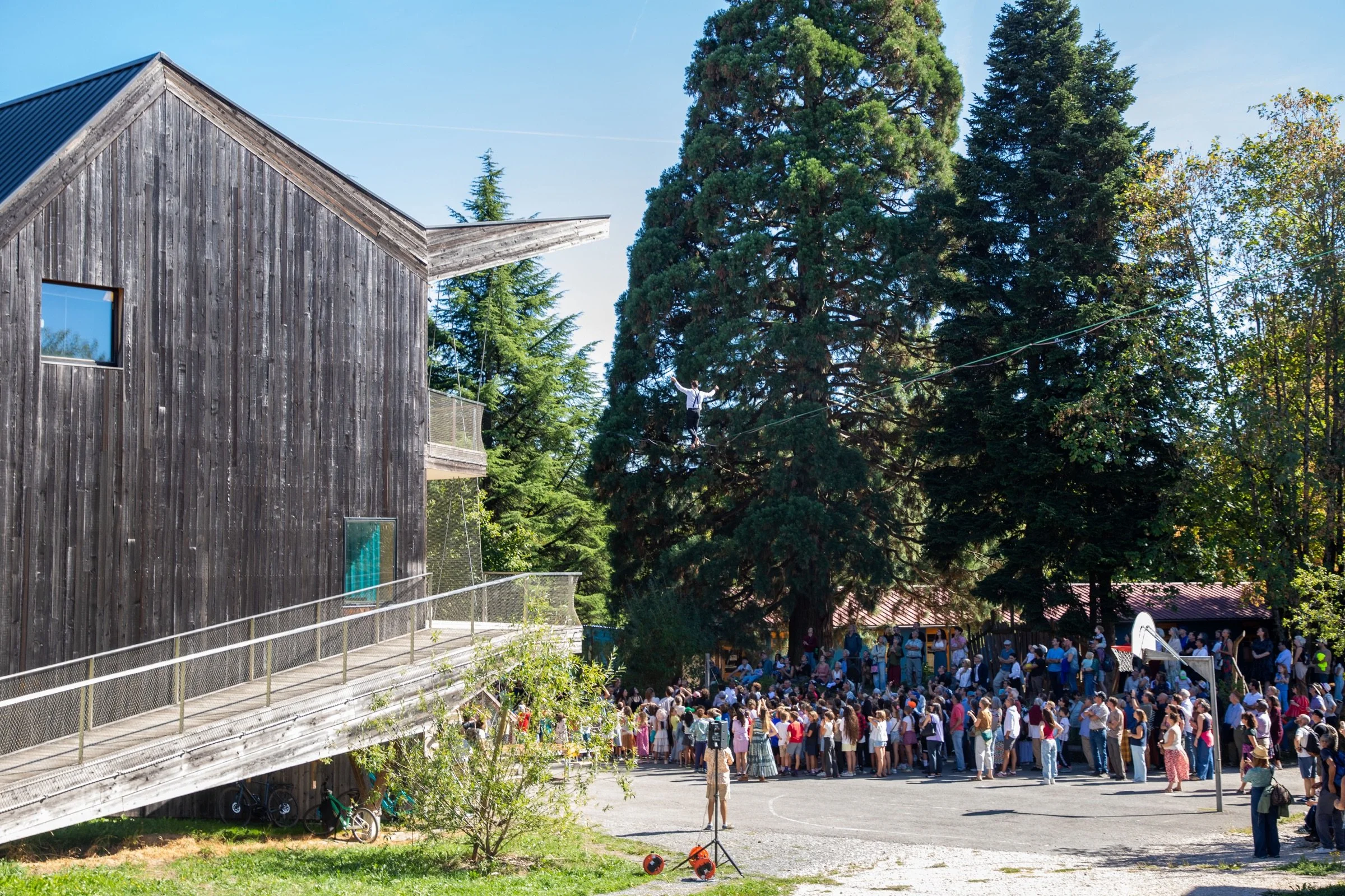Public rassemblé dans la cour de l'École Steiner à Crissier pendant un spectacle de highline, entre un bâtiment en bois et de grands conifères
