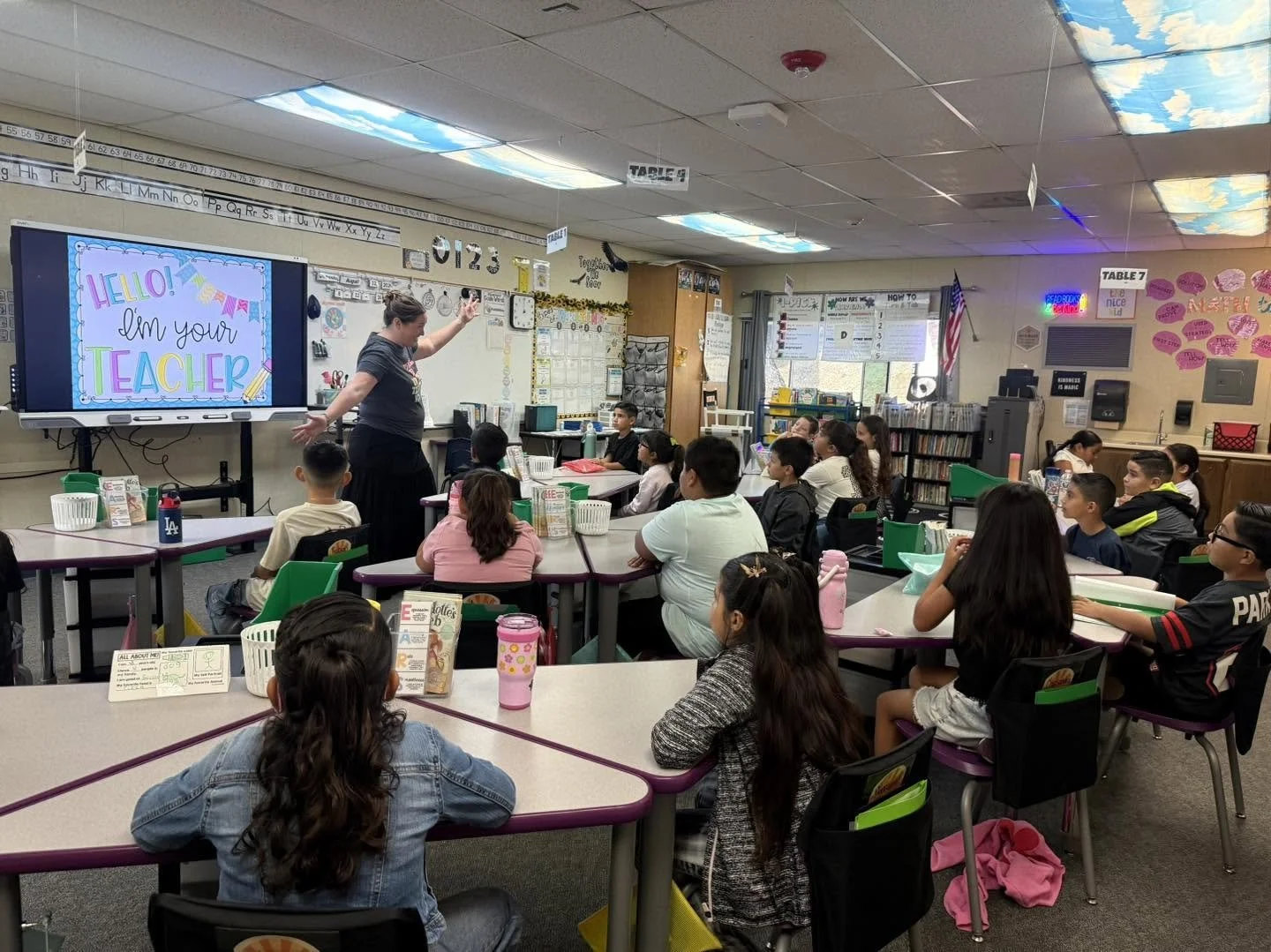 A teacher introduces himself to students seated at classroom tables.