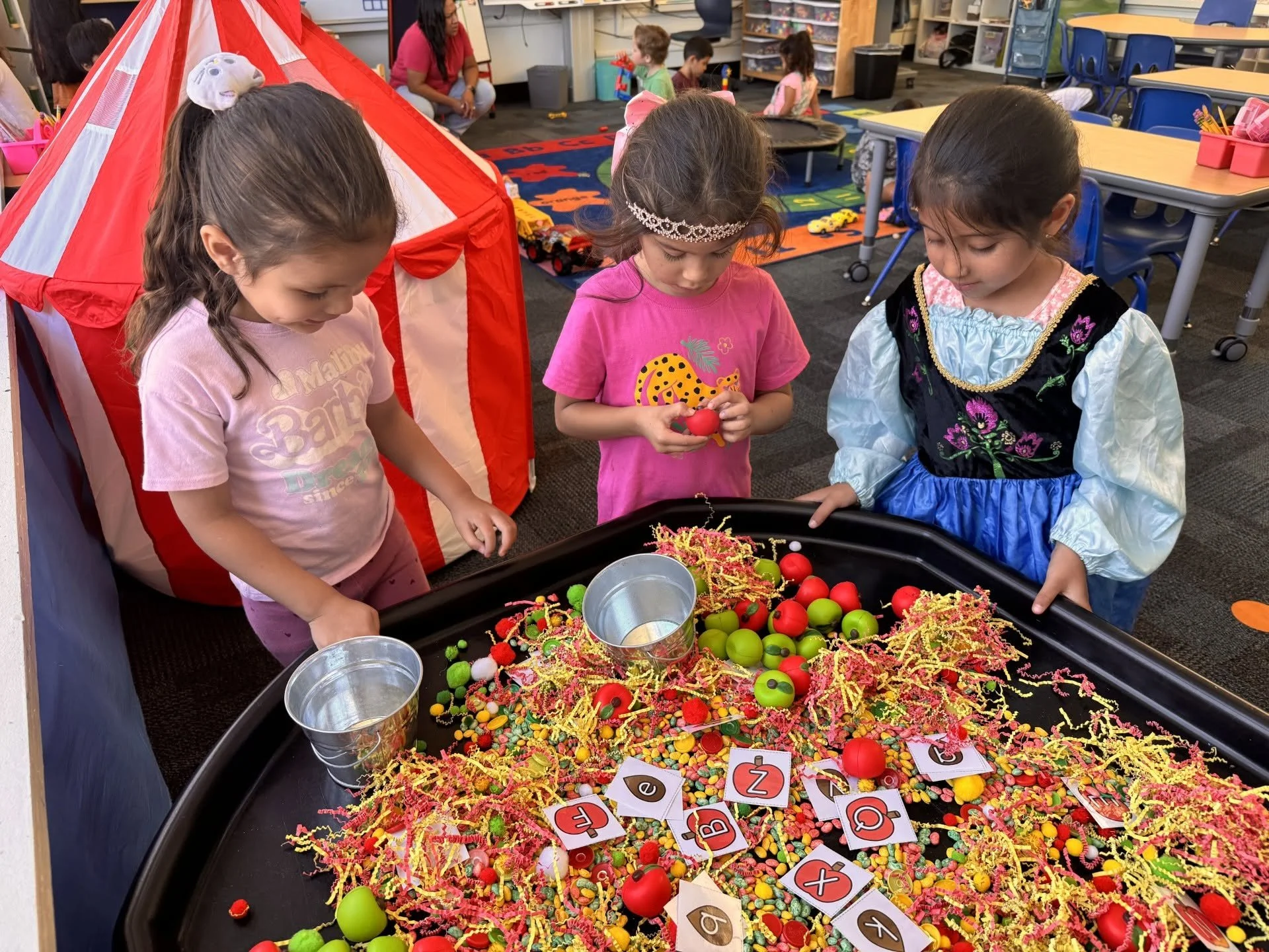 Three students explore a colorful sensory bin with toy apples and letter cards during free-choice time.