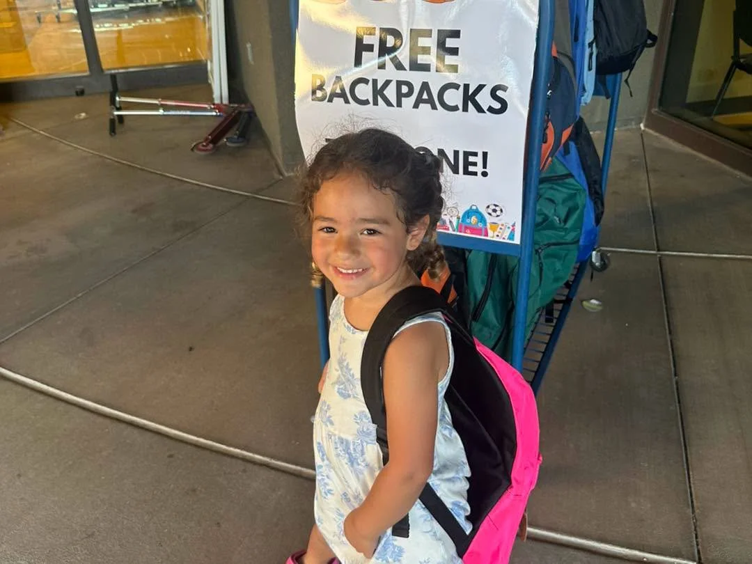 A smiling child wears a backpack beside the free backpack station.