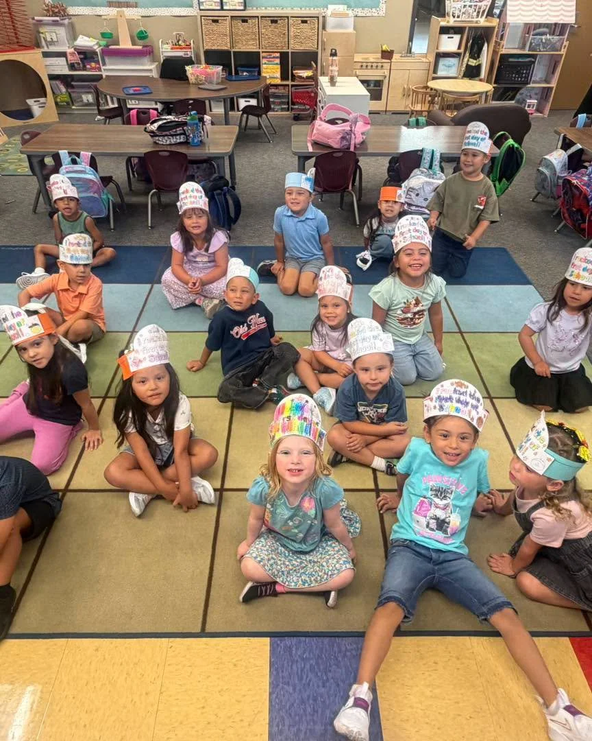 Young students wear decorated paper crowns while seated on the classroom rug.