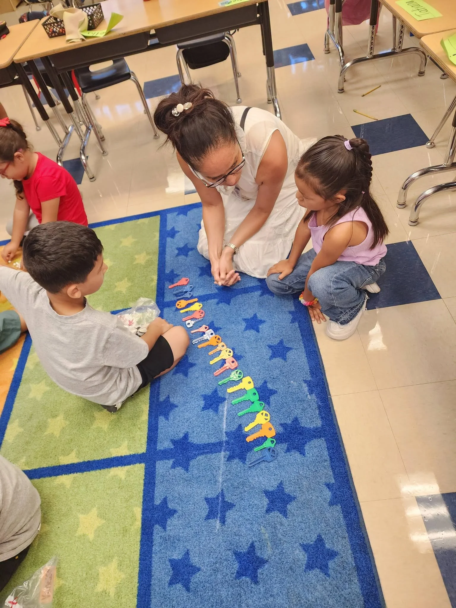 A teacher helps kindergarten students count a long row of colorful plastic keys on the classroom rug.