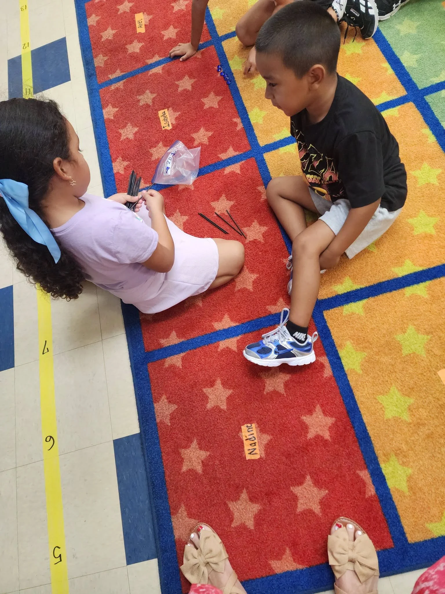 Kindergarten partners count thin black sticks together while seated on the classroom rug.
