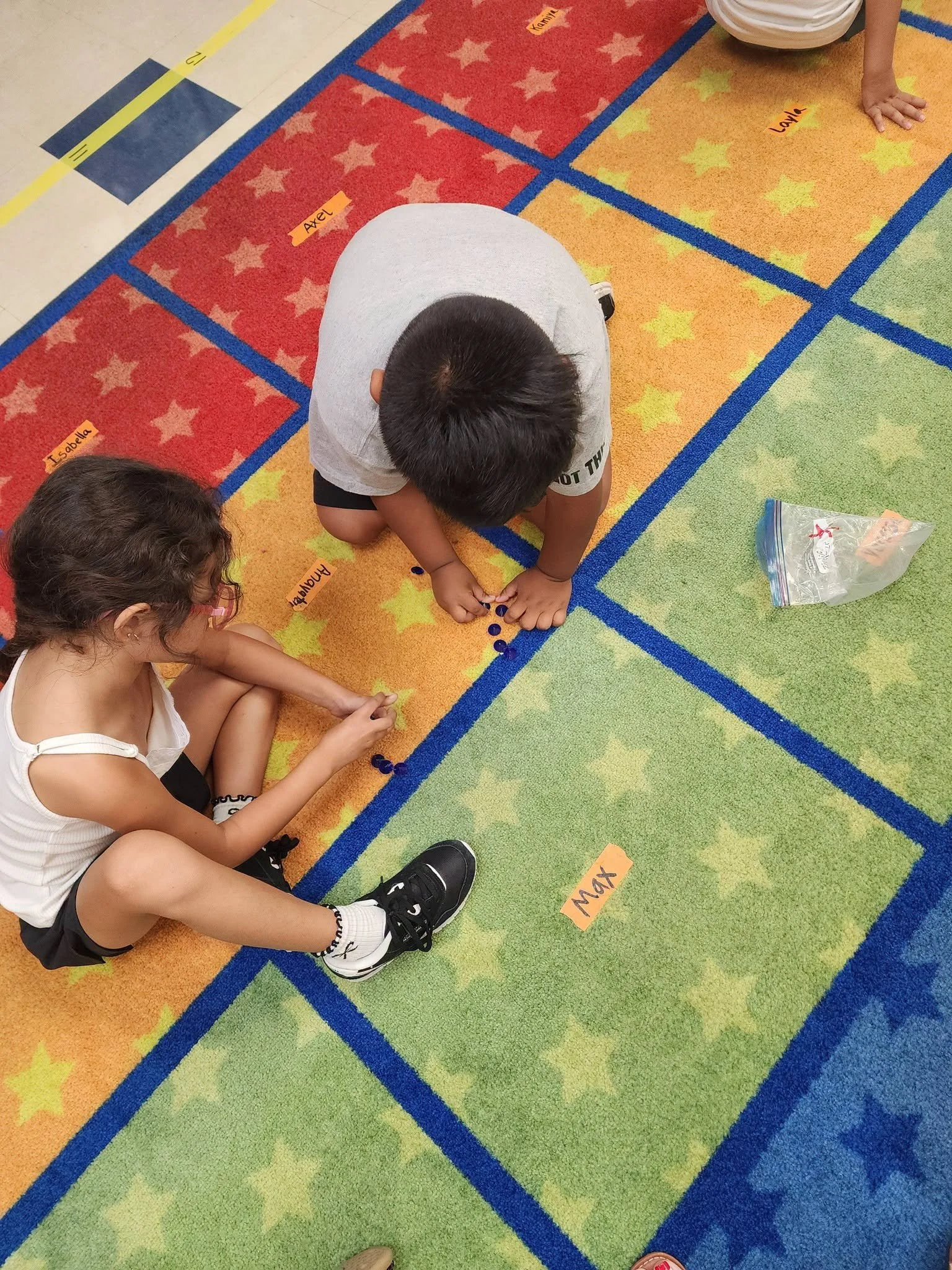 Two kindergarten students arrange small blue counters on the classroom rug.