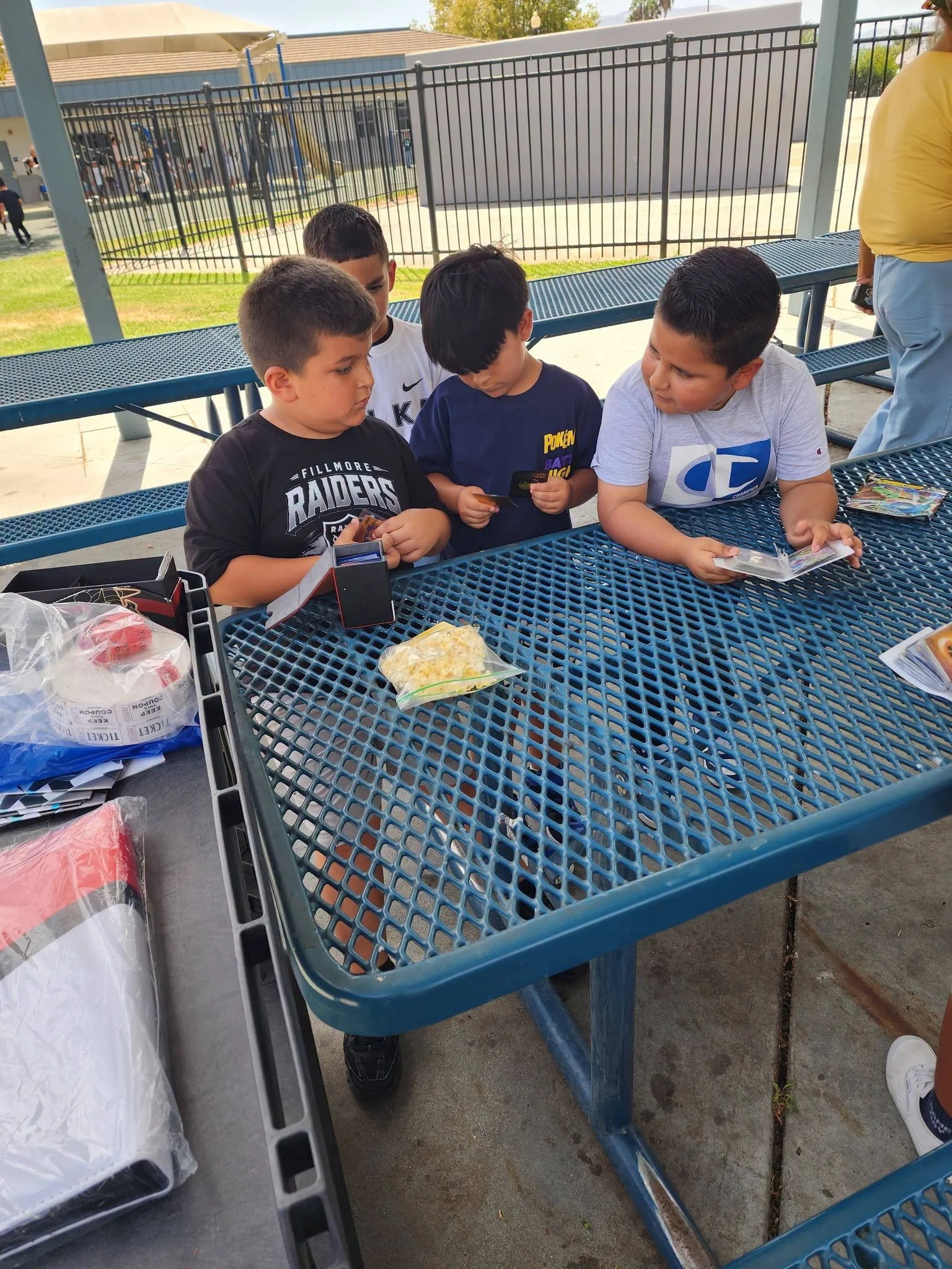 Classmates compare Pokémon cards while gathered at a shaded picnic table.
