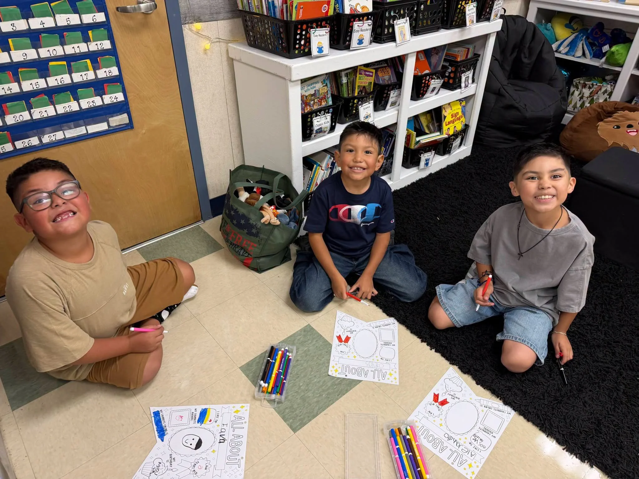 Three students work on All About Me pages beside the classroom library.