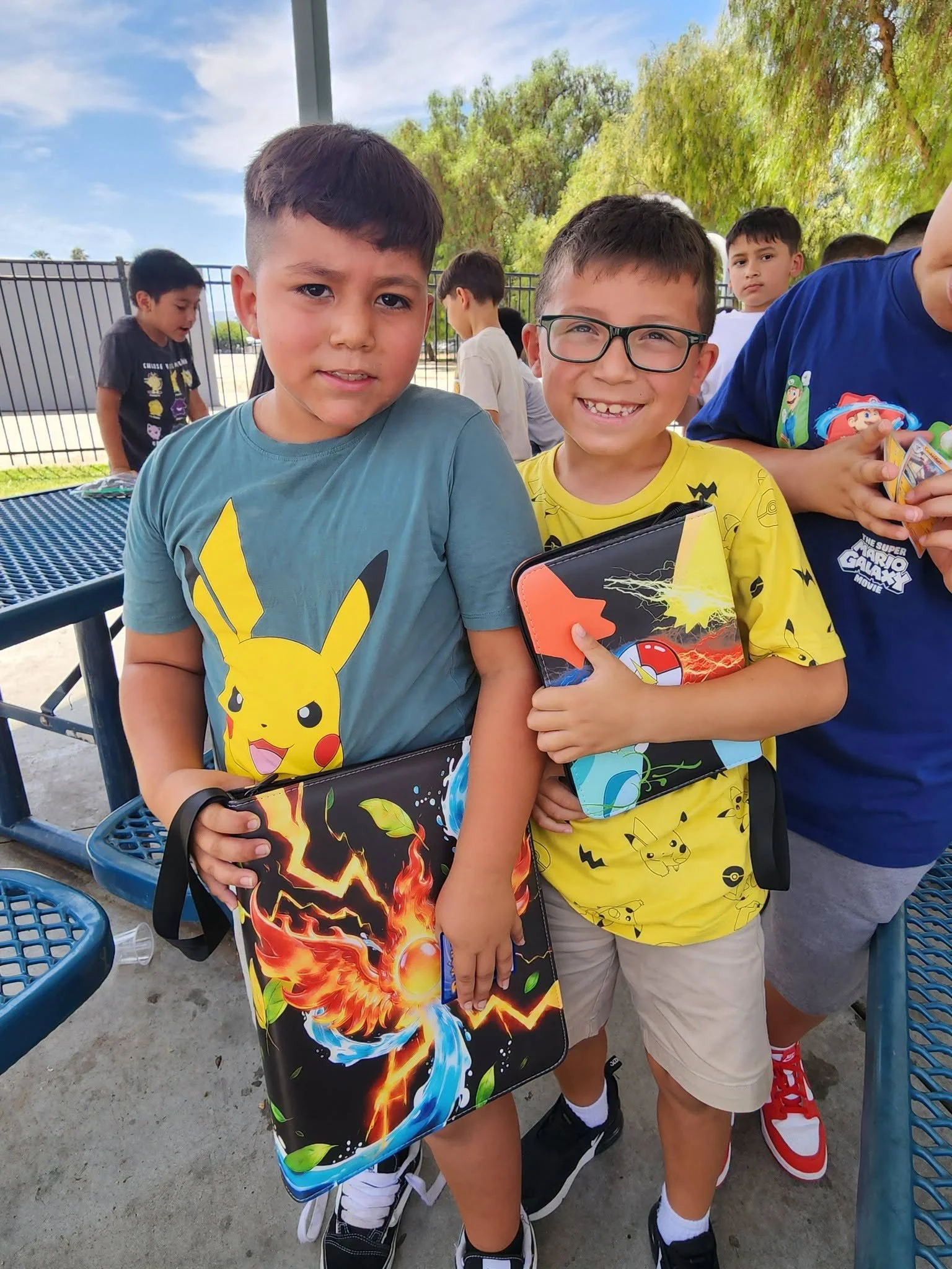 Students in Pikachu shirts hold their Pokémon card binders beside an outdoor picnic table.