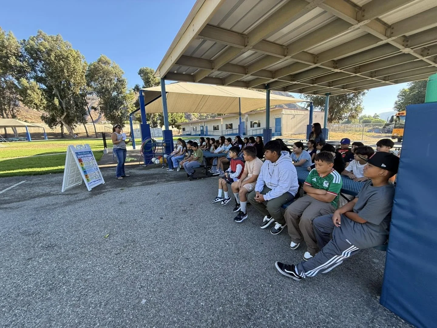 Students listen to a staff member at a shaded outdoor expectation station.