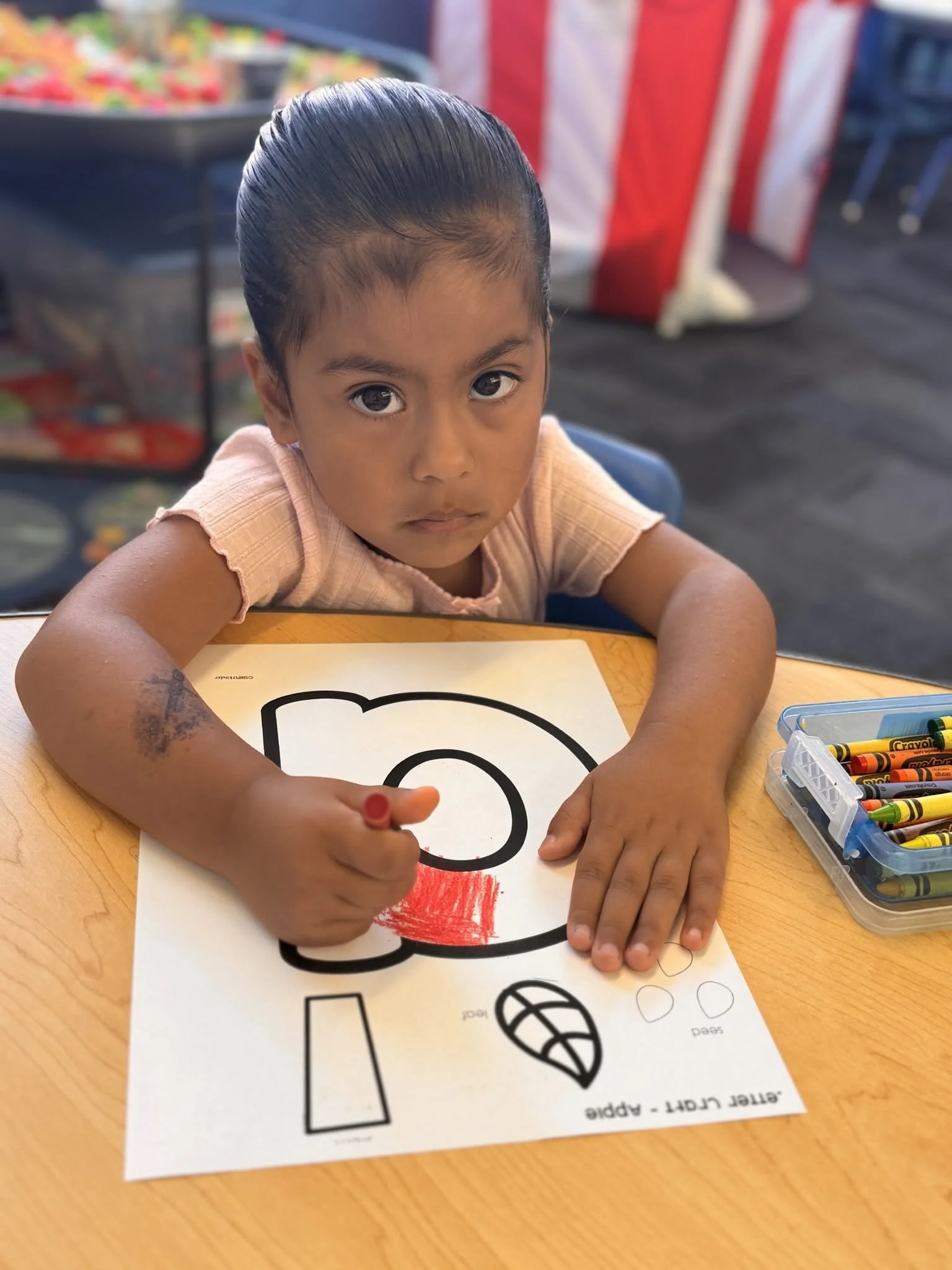 Student coloring a lowercase letter a worksheet at a classroom table.