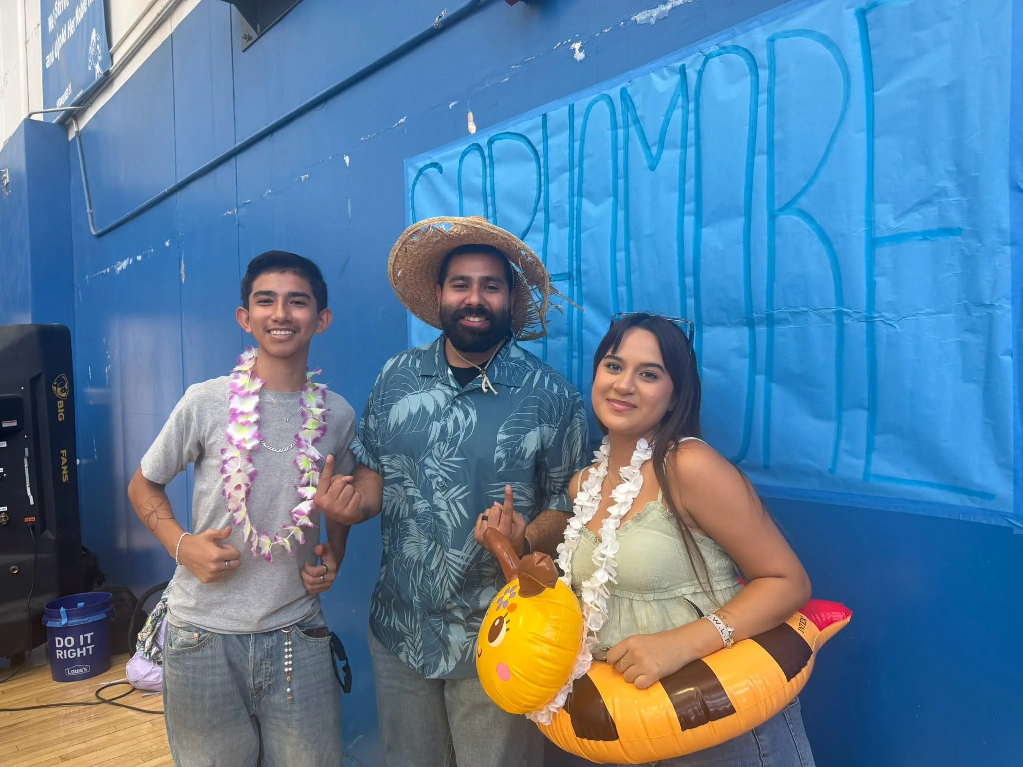 Three people in tropical-themed spirit wear pose beside a sophomore banner in the gym.