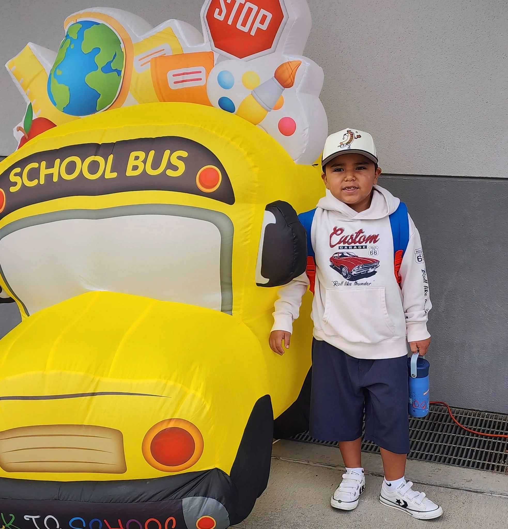 A student with a cap and backpack poses beside the yellow school bus.