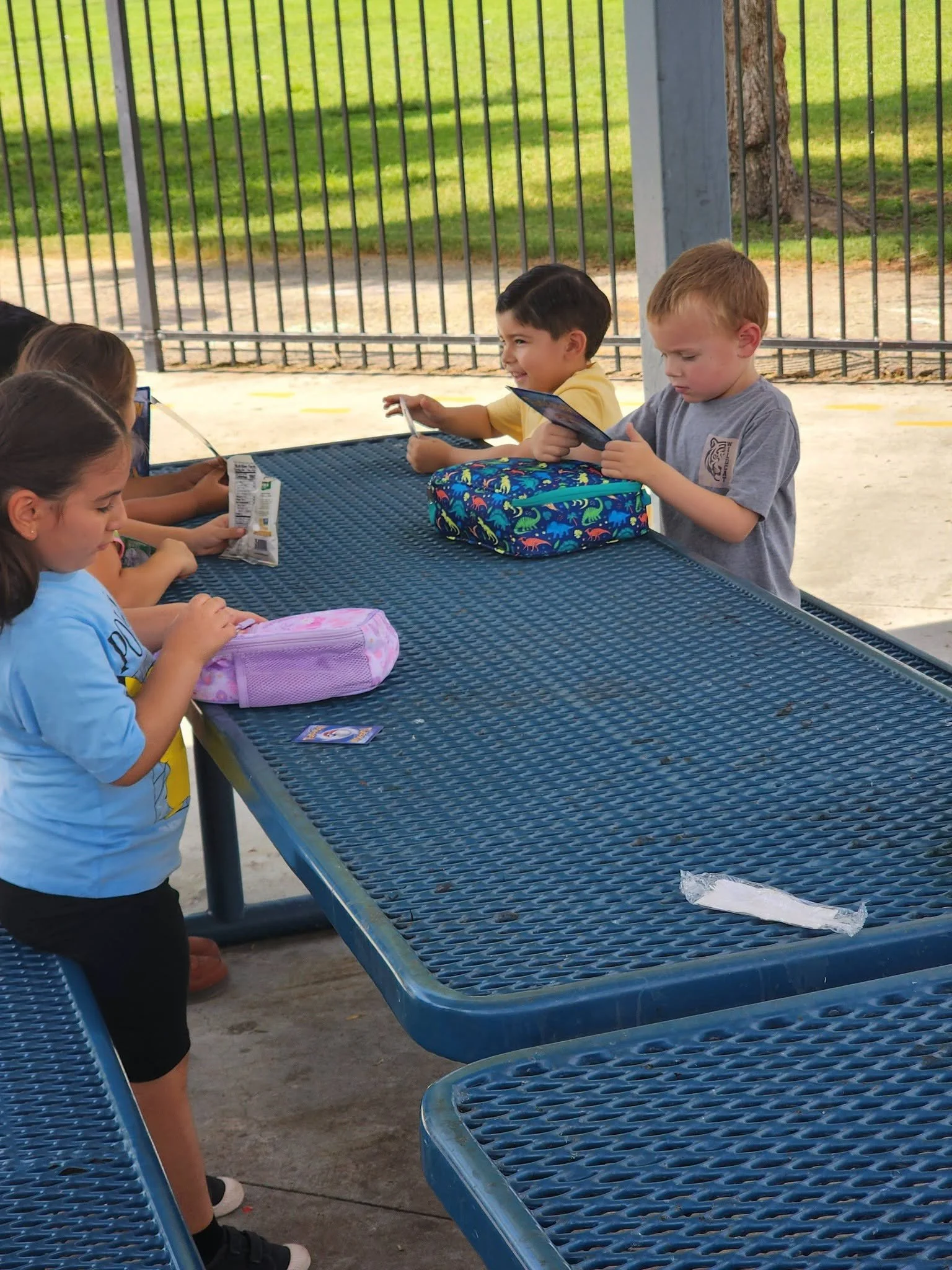 Students take out Pokémon cards and card cases at an outdoor lunch table.