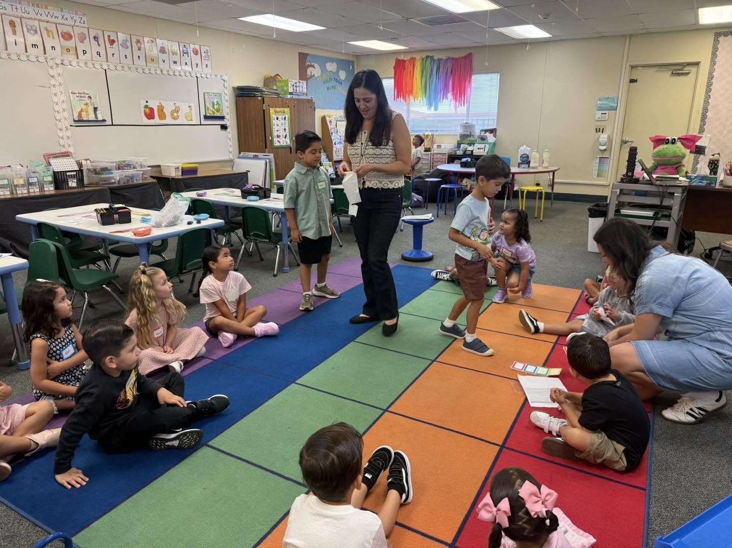 Students and staff take part in a classroom activity on a colorful rug.