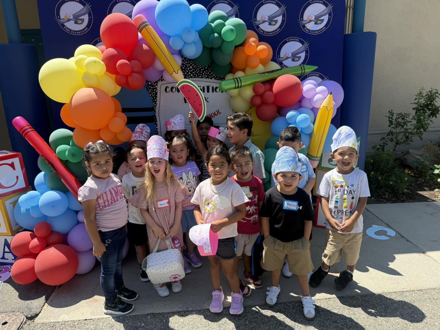 Young students celebrate their first day beside colorful balloons and school supplies.