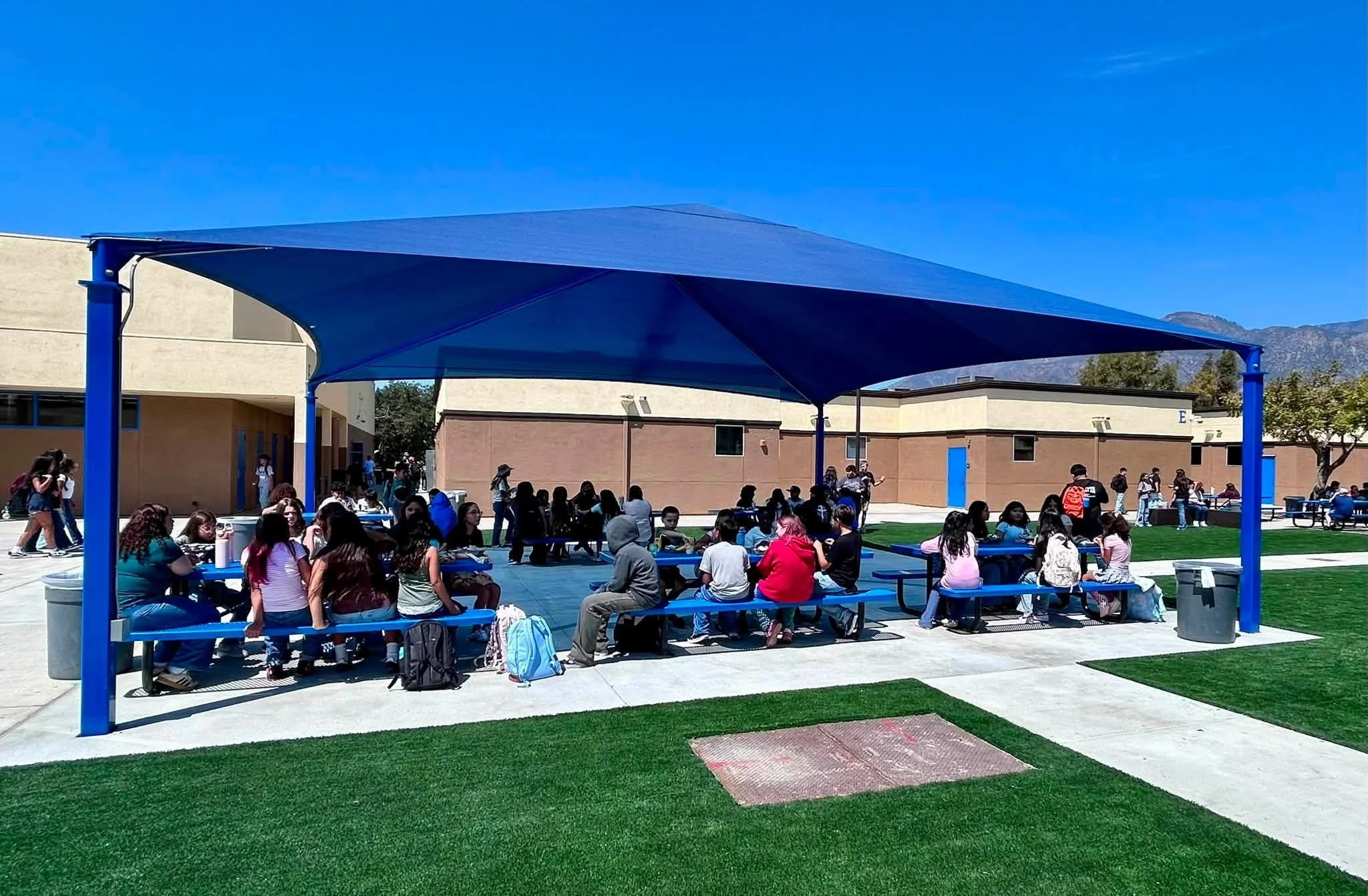 Students gather at picnic tables beneath a new blue shade canopy.