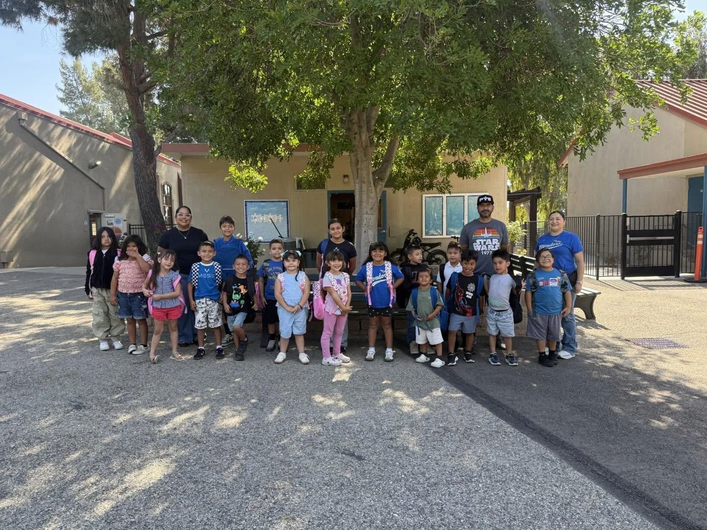Piru students show their backpacks while standing with adults beneath a tree.
