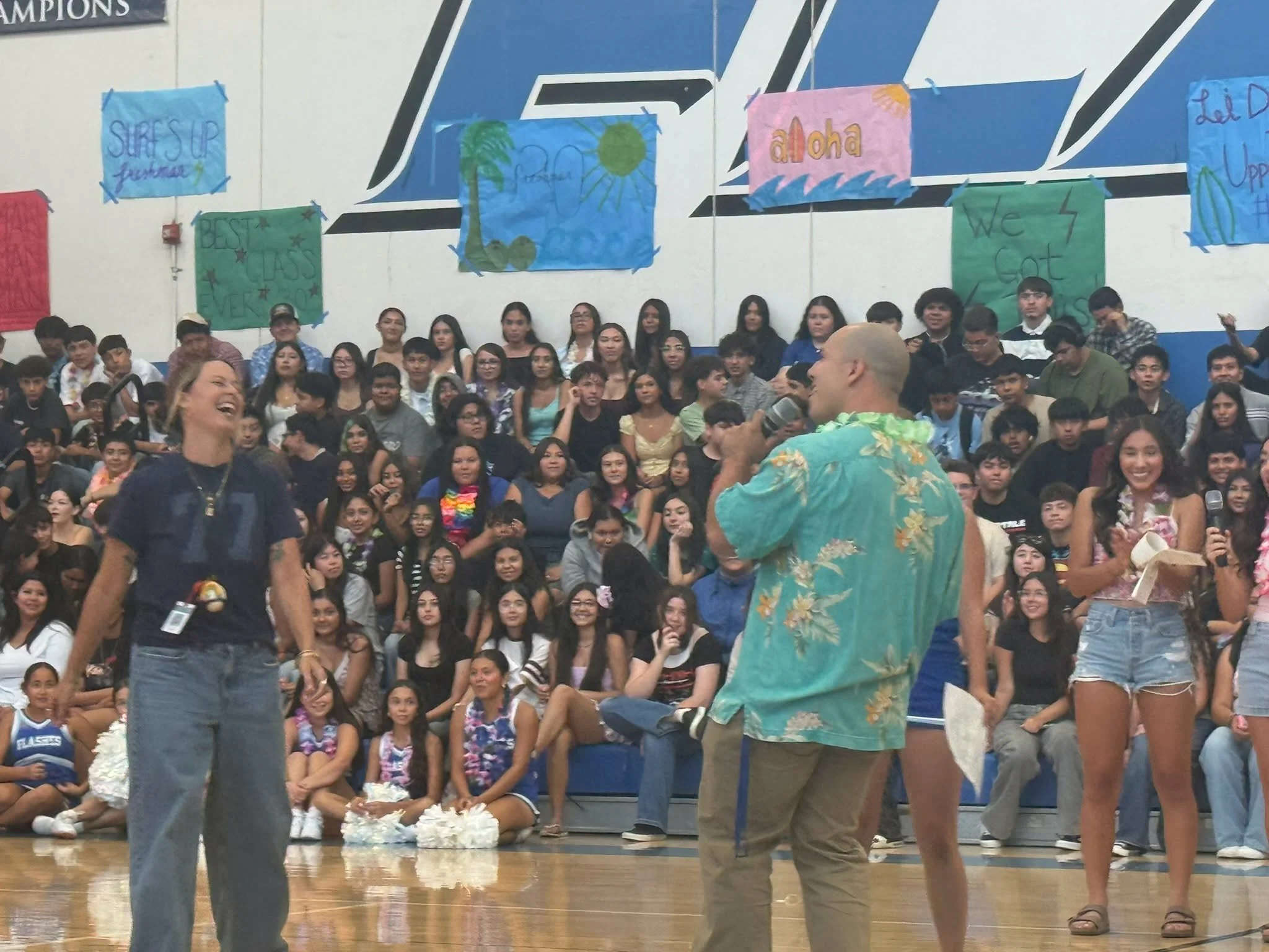 Assembly hosts speak with microphones in front of students filling the Flashes gym bleachers.