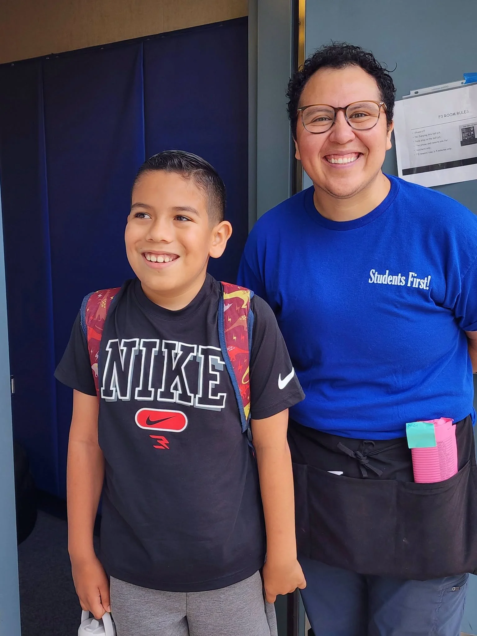 A staff member and student smile together outside a classroom.