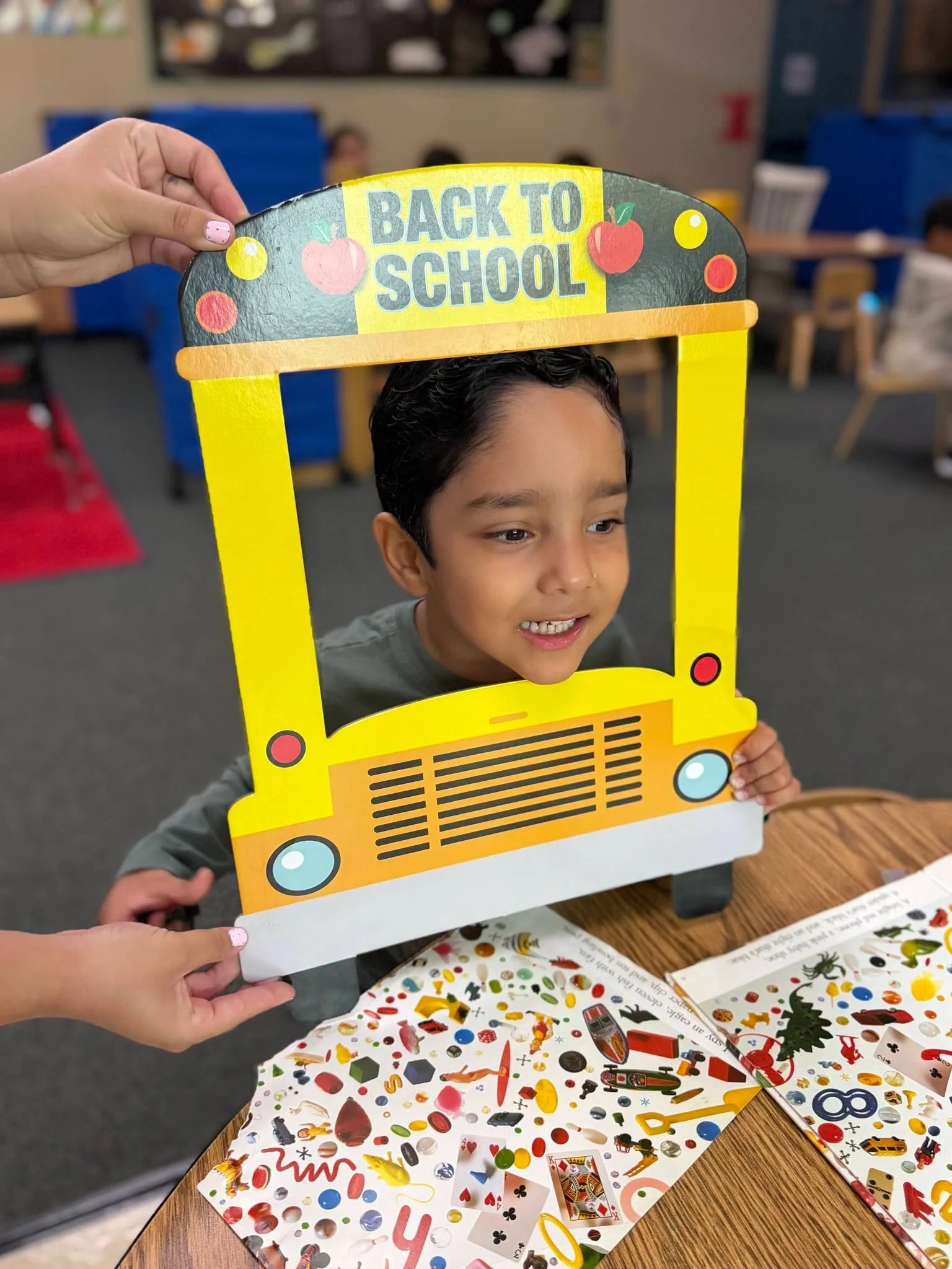 A student smiles through a school-bus photo frame at a classroom table.