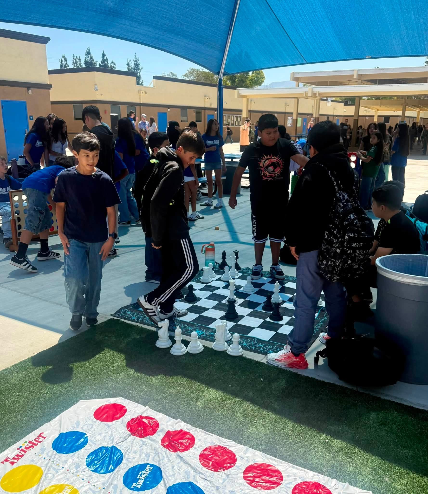 Students play oversized chess beside a Twister mat under the shade canopy.