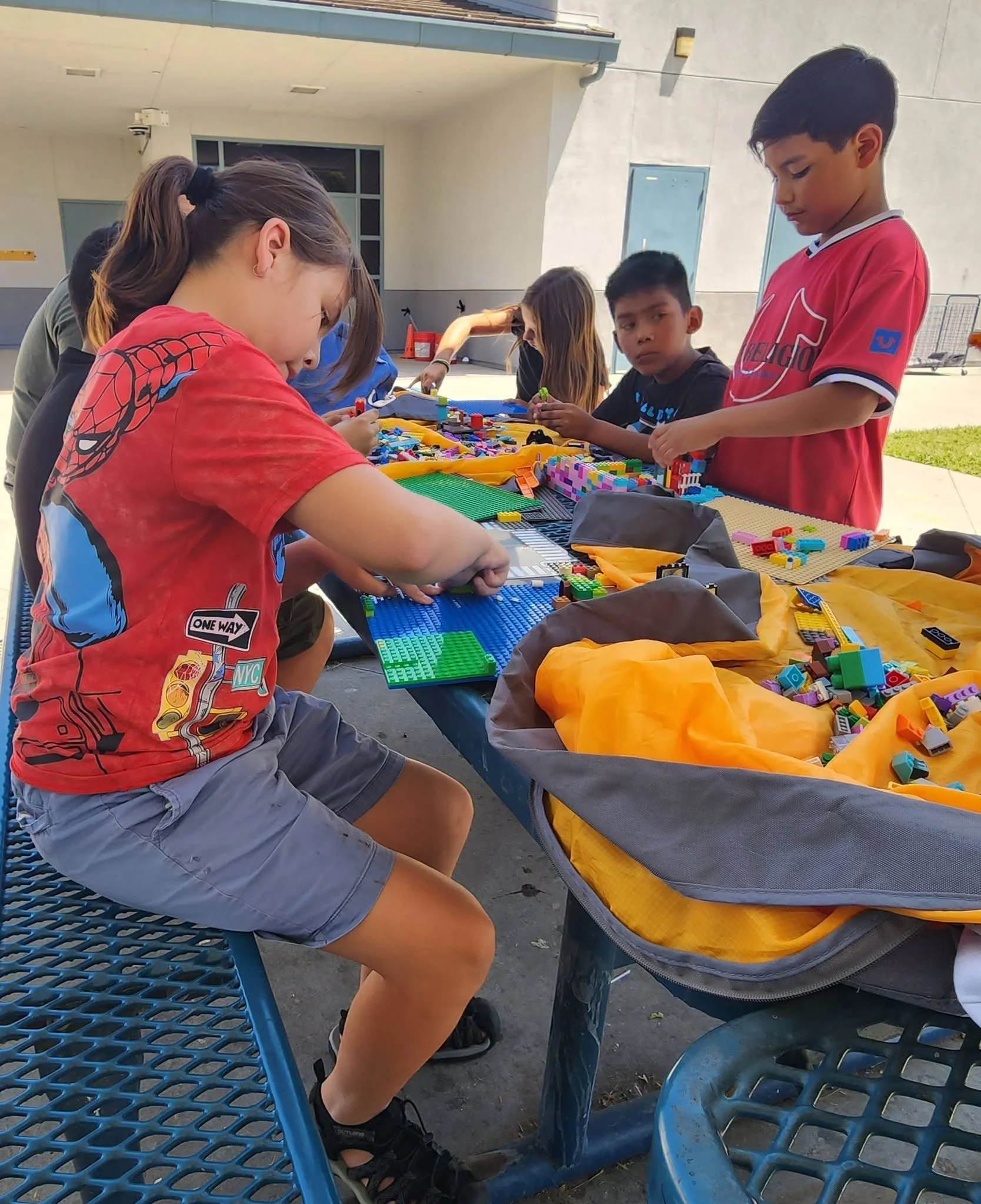 Students in red shirts build with Lego bricks at an outdoor picnic table.