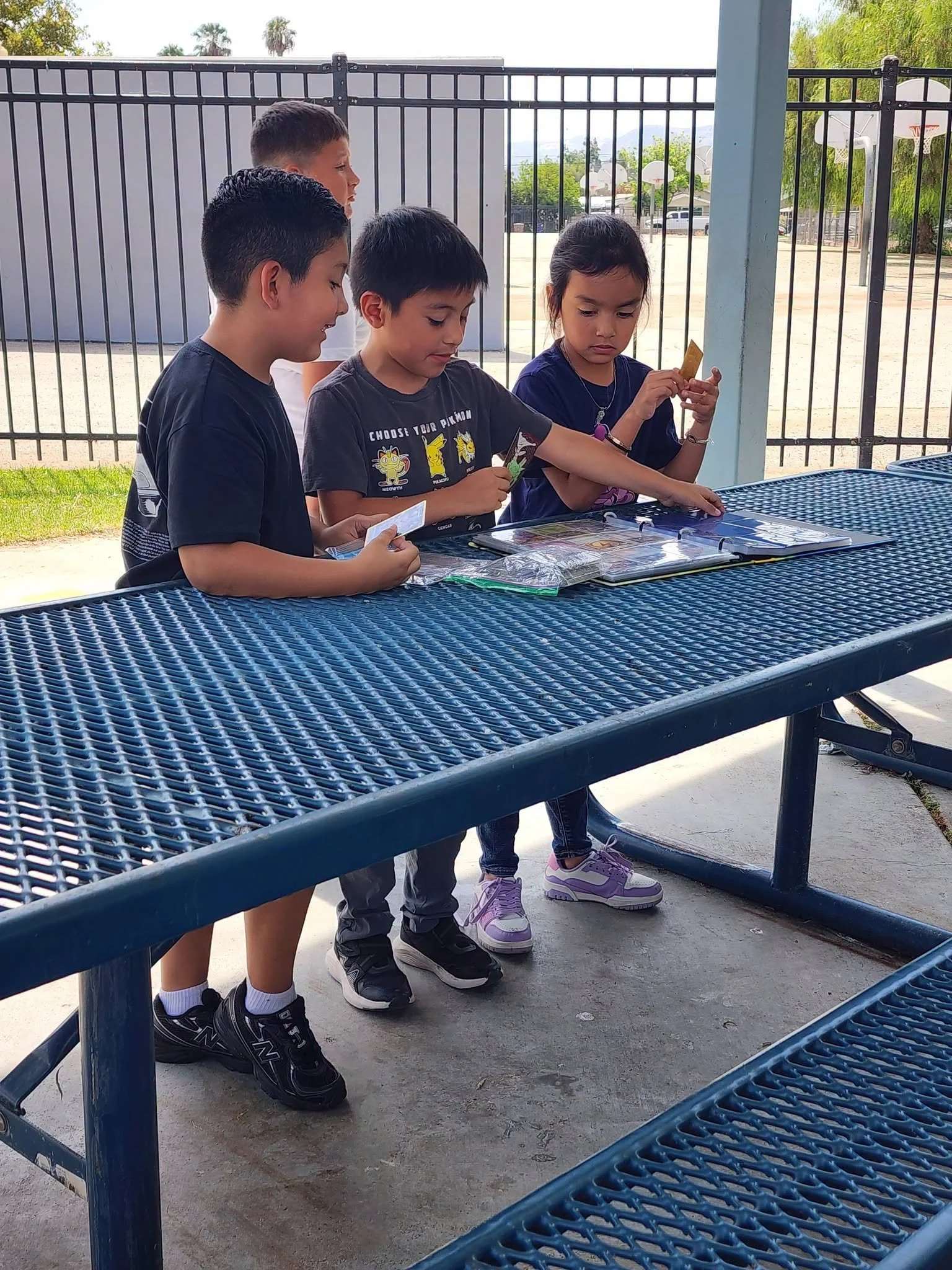 Students discuss Pokémon cards around an open binder on a blue picnic table.