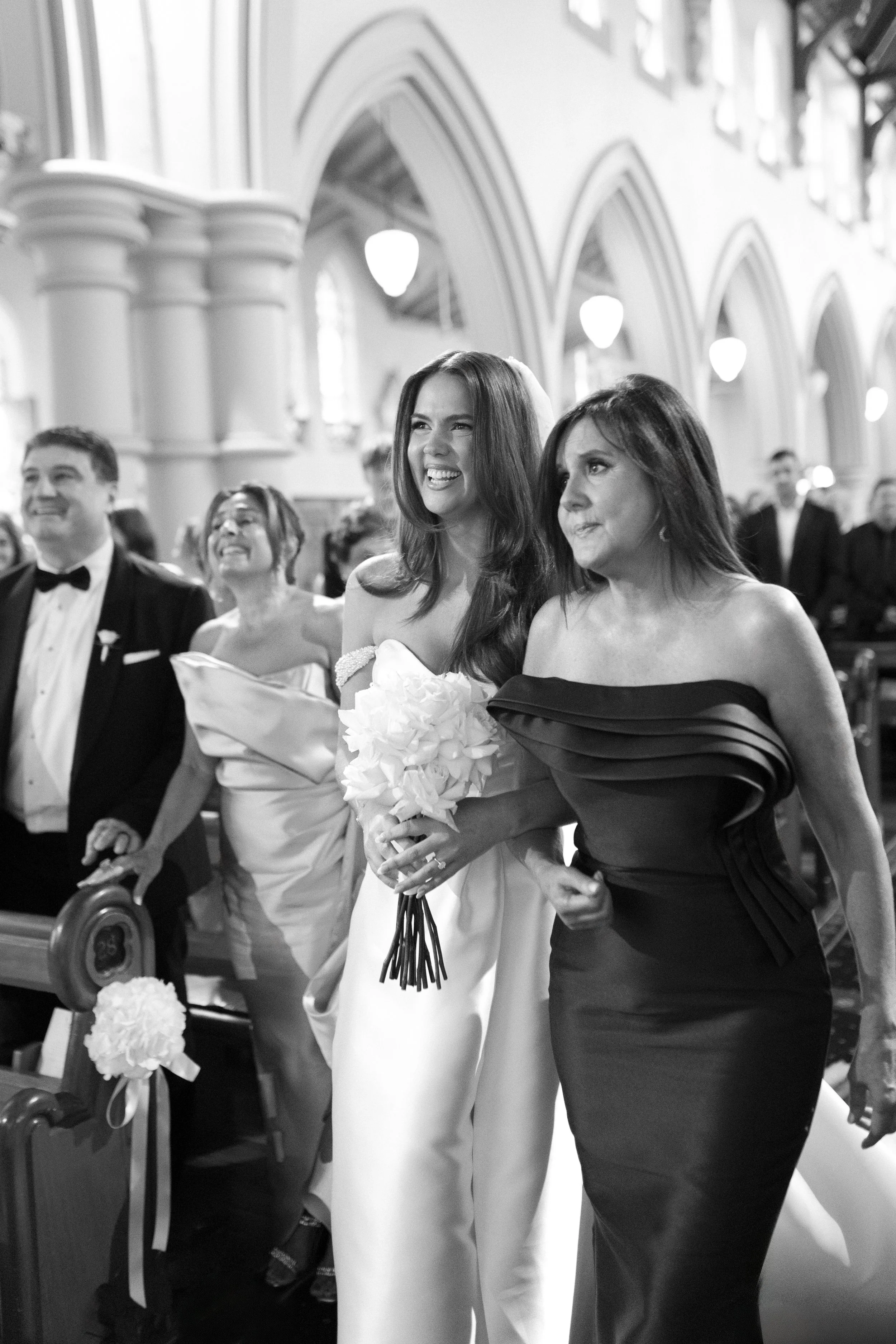 A joyful bride holding a bouquet of white flowers is surrounded by guests in a church during a wedding ceremony, with smiling faces and formal attire.